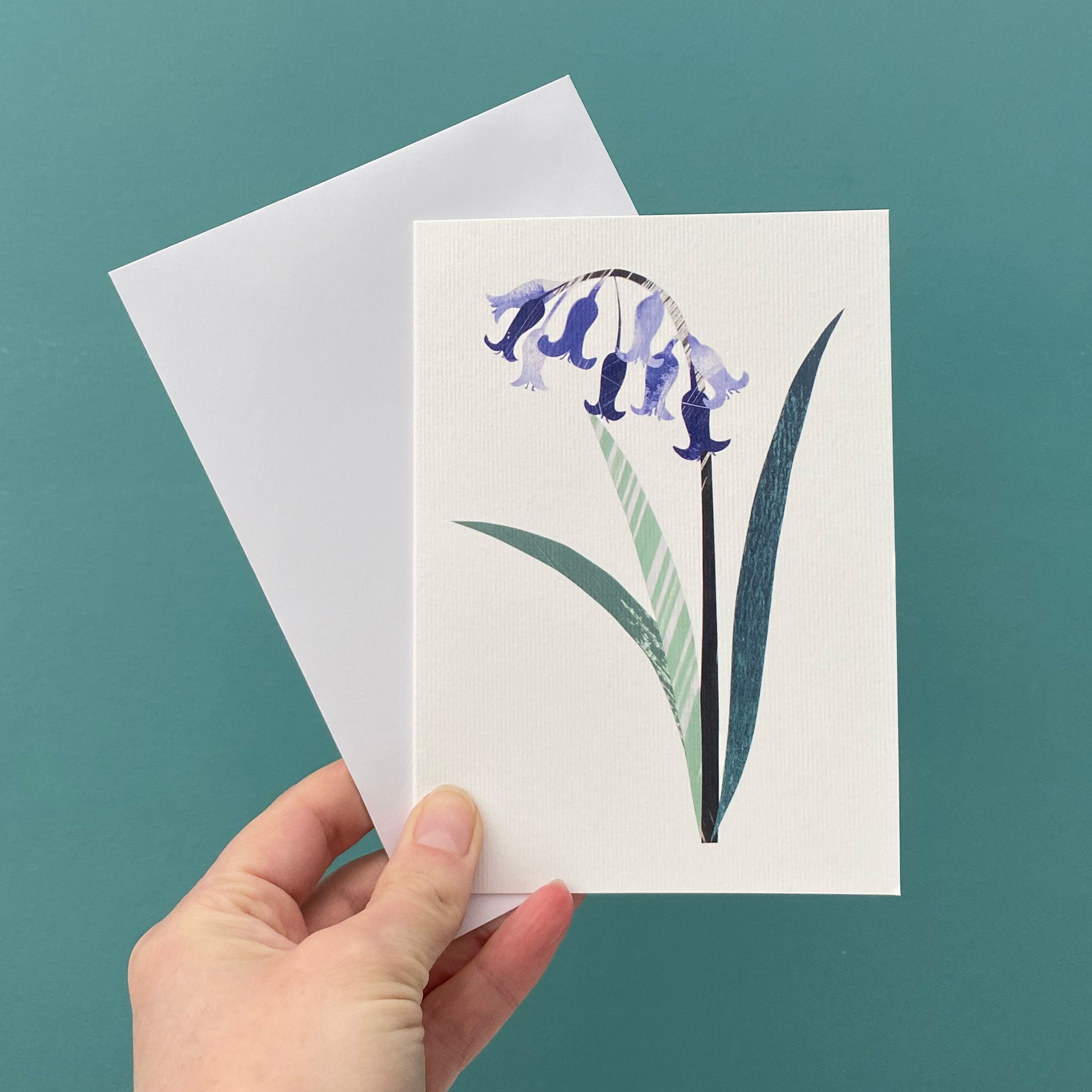 A card featuring a single Bluebell flower with green leaves on a white background is being held with a white envelope against a blue green background.