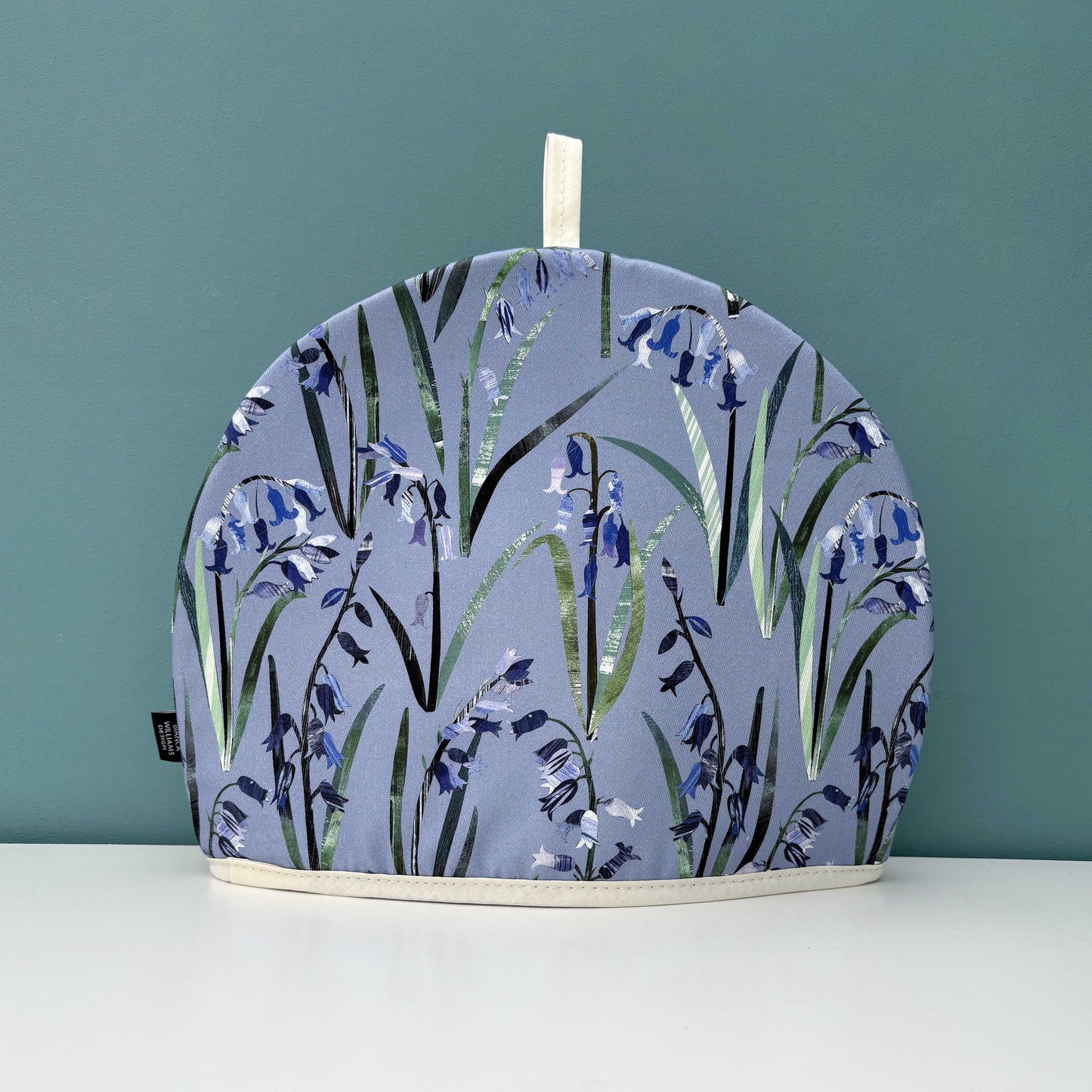 A Bluebell Tea cosy with a Brand label and a hanging loop at the top has been placed  on a white surface against a green background.