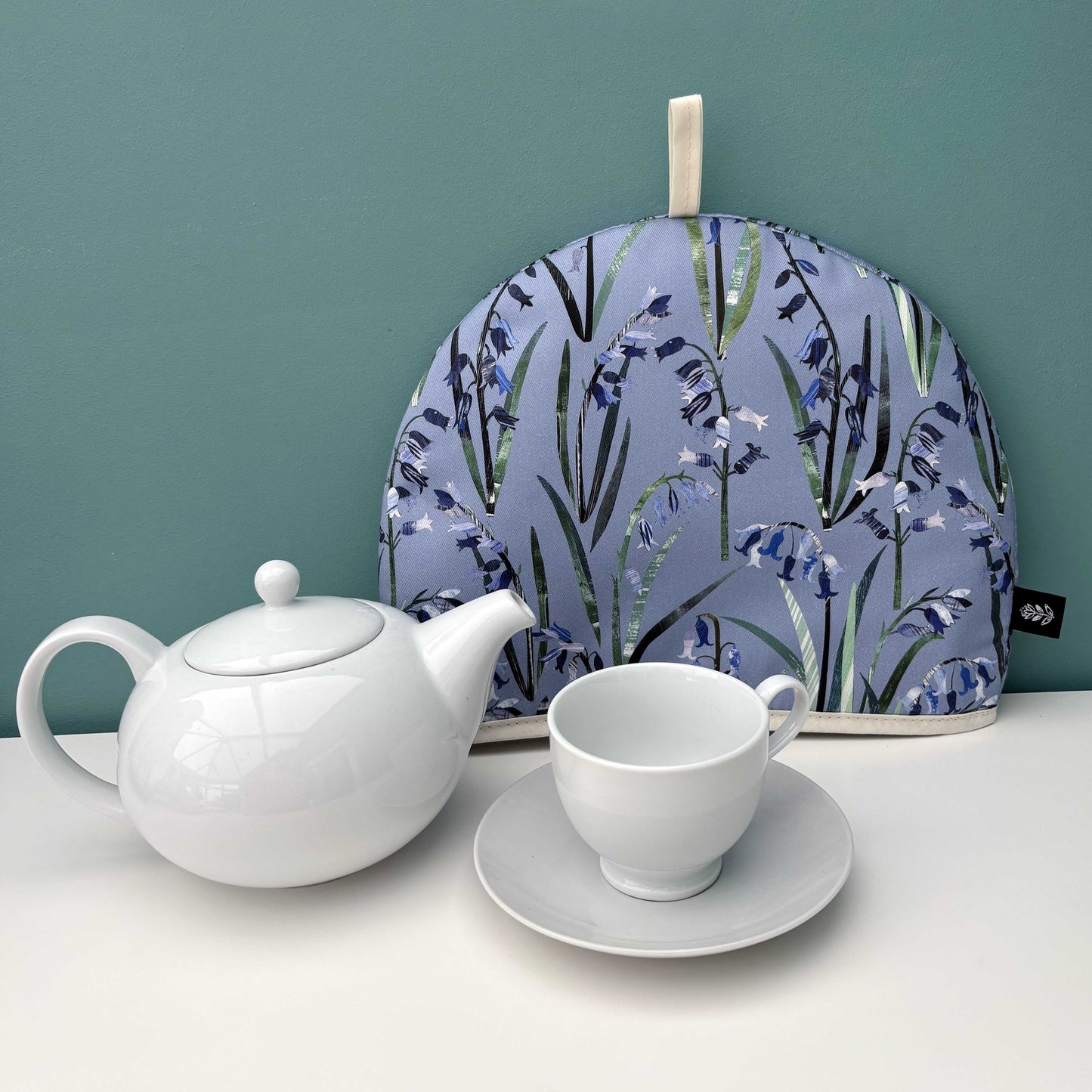 A Bluebell Tea Cosy and a white teapot, cup, and saucer on a white surface against a teal background.