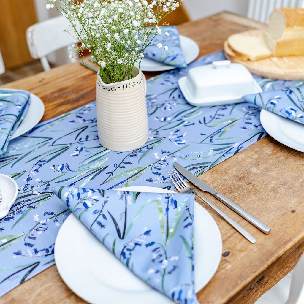 Dining table setting with bluebell floral table runner and matching napkins, white plates, and a vase of flowers.