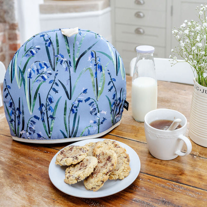 A Bluebell Tea Cosy has been placed on a wooden kitchen table with some cookies a cup of tea and a jug of flowers.