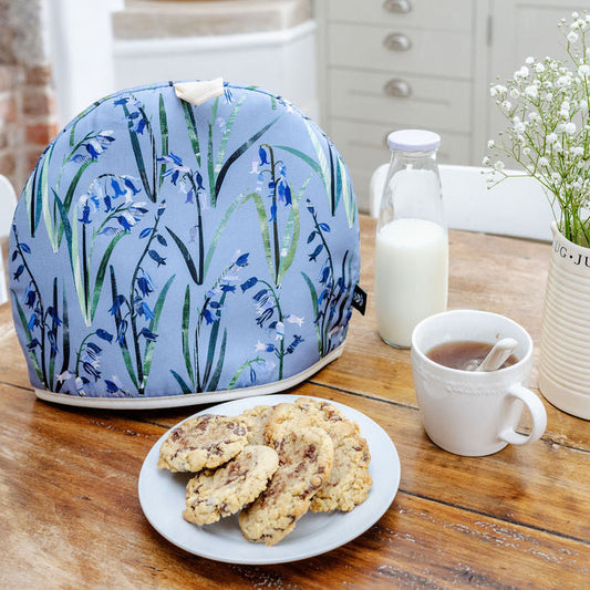 A Bluebell Tea Cosy has been placed on a wooden kitchen table with some cookies a cup of tea and a jug of flowers.