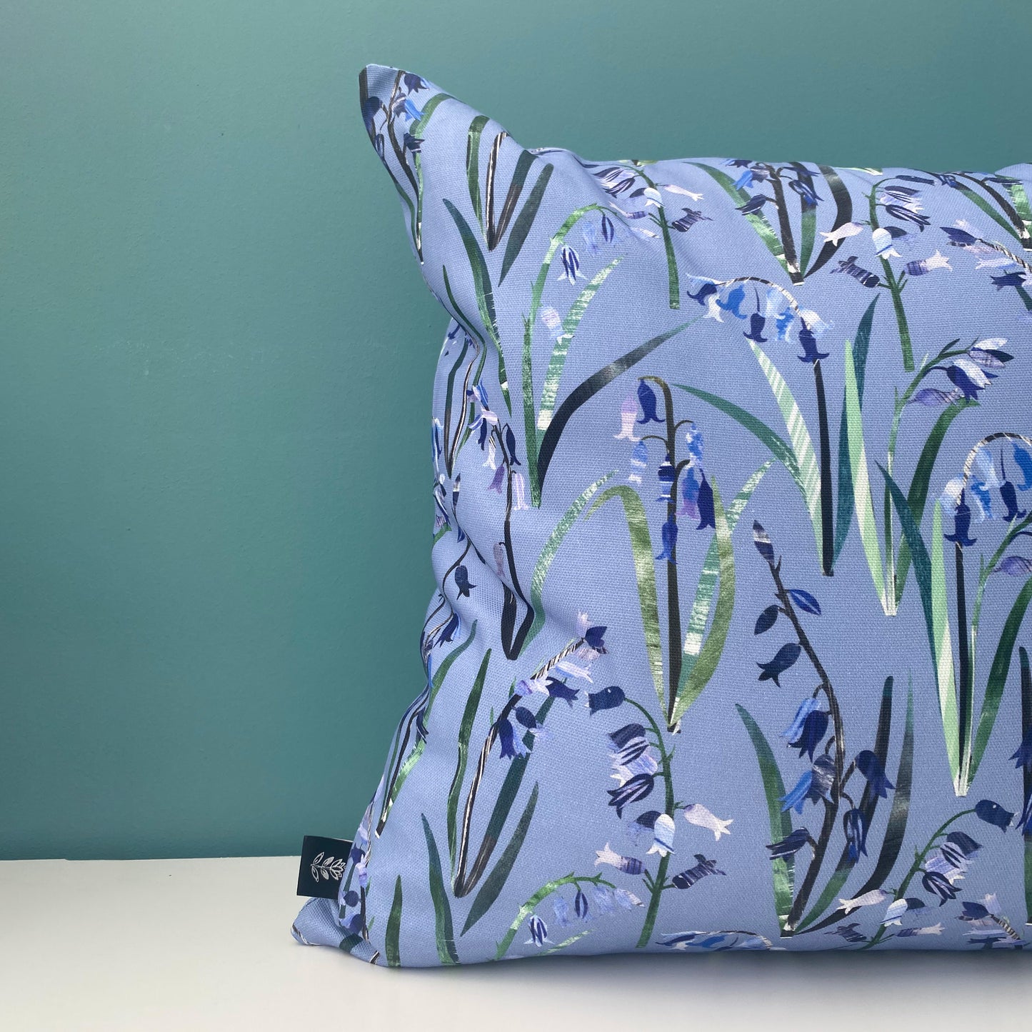 A close up of a Blue Cushion featuring pretty textured Bluebell Flowers with green leaves has been printed onto a Cornflower Blue background. The cushion has been placed onto a white shelf with a blue/green wall behind.