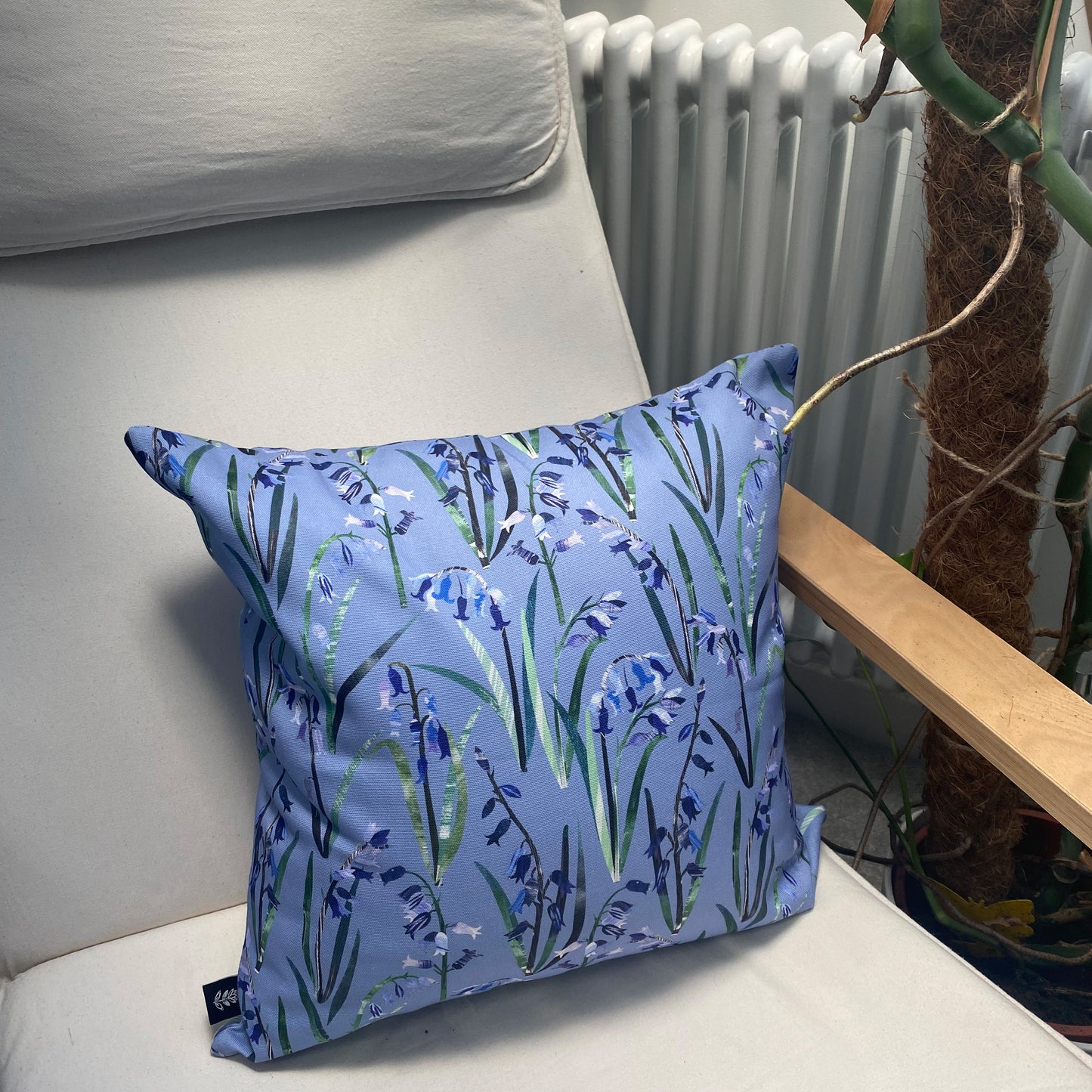 A Bluebell Cushion has been placed onto a cream canvas and wooden armchair, you can see a radiator and plant in the background.