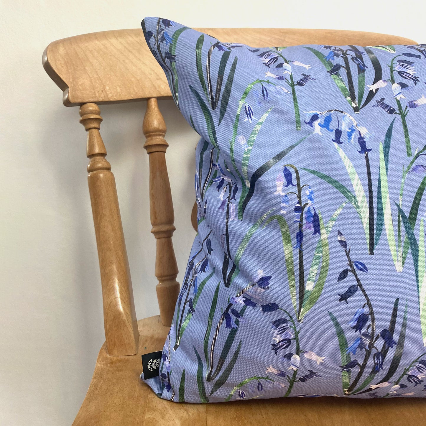 A Bluebell Cushion has been placed onto a wooden Dining chair against a pale grey wall.