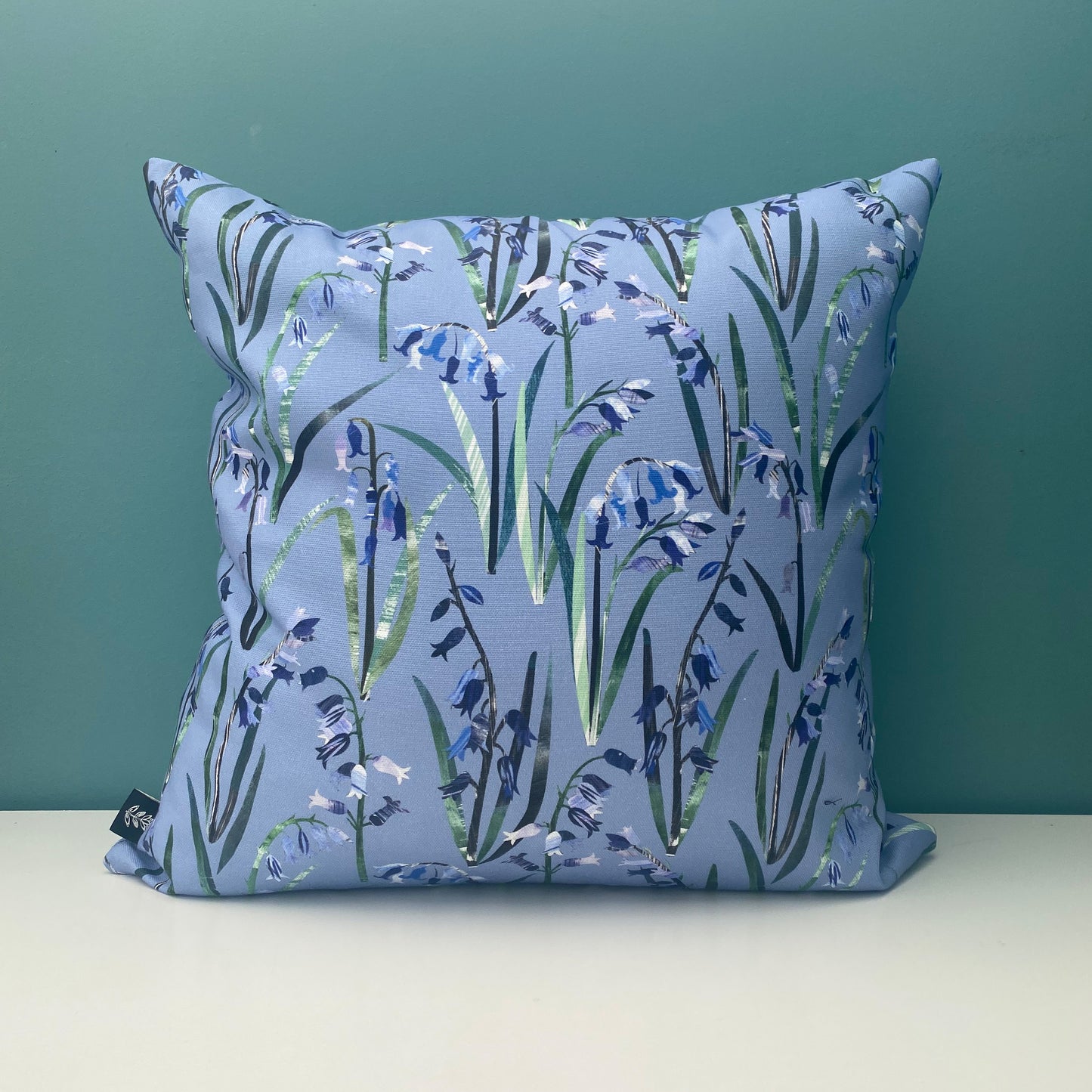 A Bluebell Cushion featuring pretty textured Bluebell Flowers with green leaves printed onto a cornflower blue background. The cushion has been placed onto a white shelf with a blue/green wall behind.