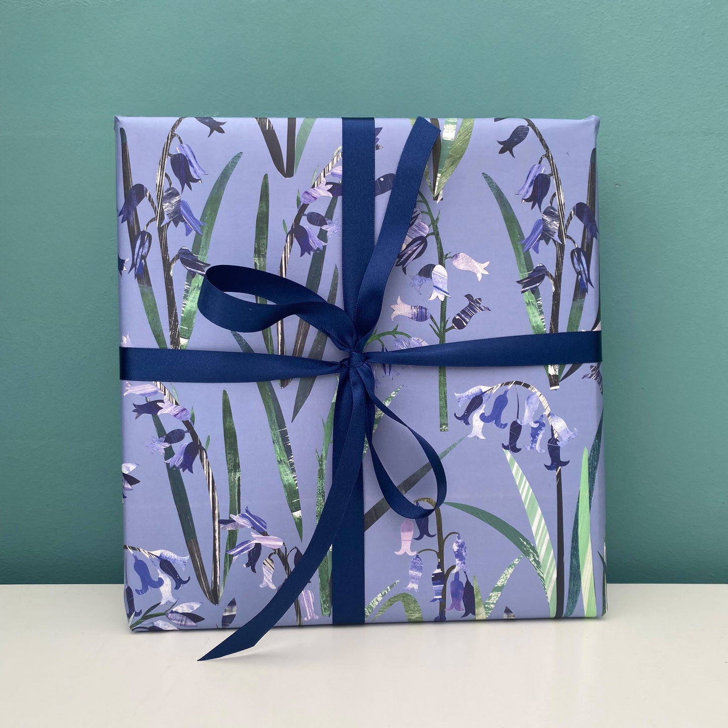 A present has been wrapped in Bluebell Gift Wrap and has been tied with a coordinating dark blue satin ribbon and placed on a white shelf against a blue green backdrop.