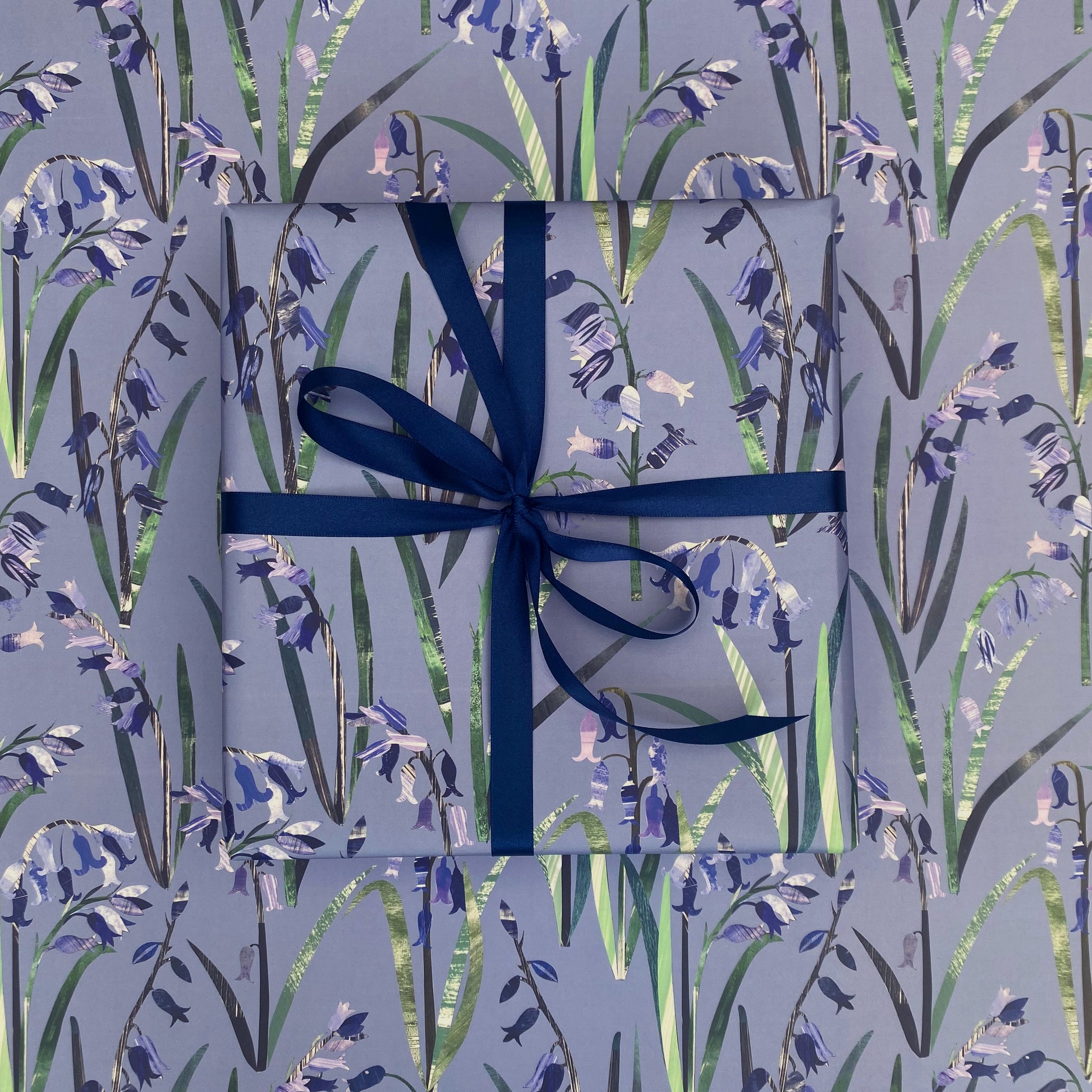 A present has been wrapped in Bluebell Gift Wrap and has been tied with a coordinating dark blue satin ribbon and placed on a sheet of laid out bluebell gift wrap. The gift wrap features Blue Bluebells with green leaves on a blue background.