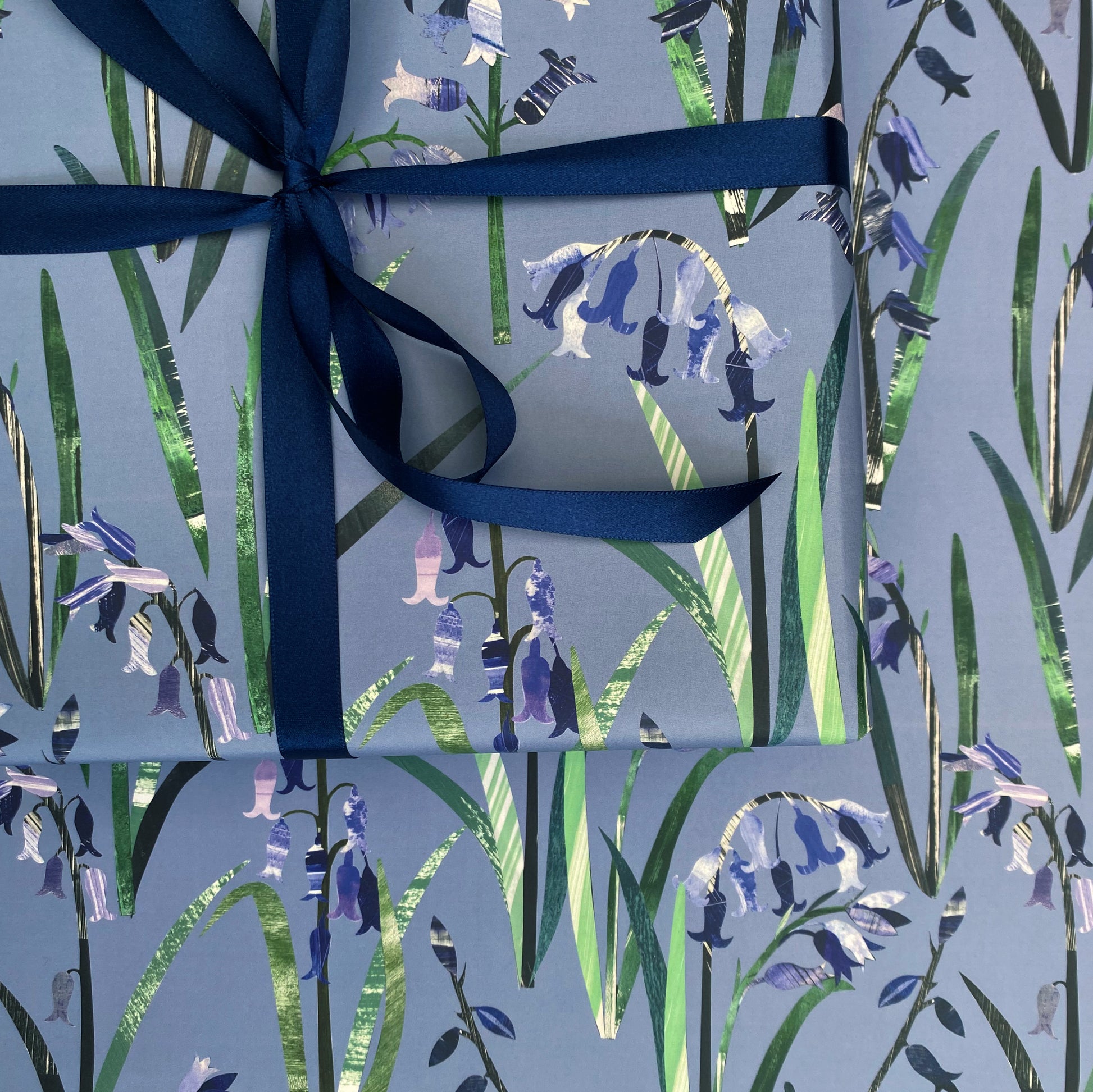 A close up of a present that has been wrapped in Bluebell Gift Wrap and tied with a coordinating dark blue satin ribbon and placed on a sheet of laid out bluebell gift wrap.