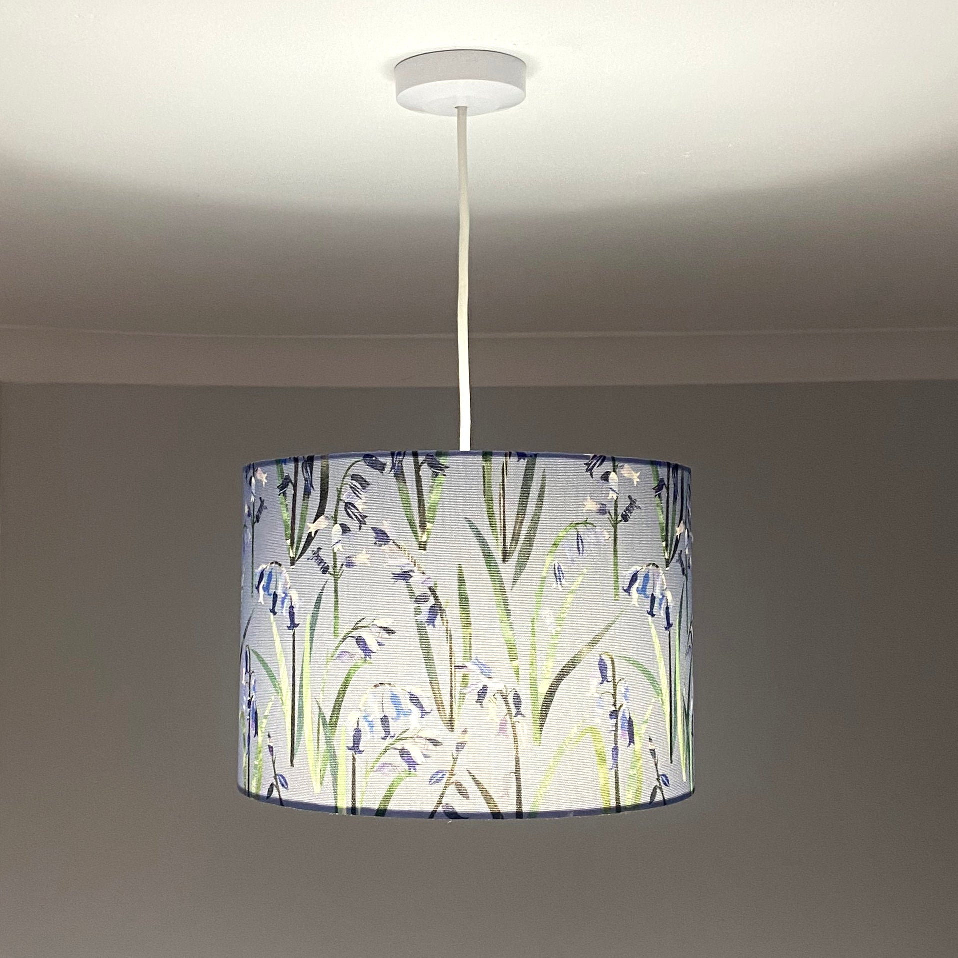 Bluebell lampshade, shown here in a Medium Size and as a Ceiling Pendant. The design features Blue Bluebell flowers with green leaves on a light blue background.  The bulb is shown switched on in this photograph.