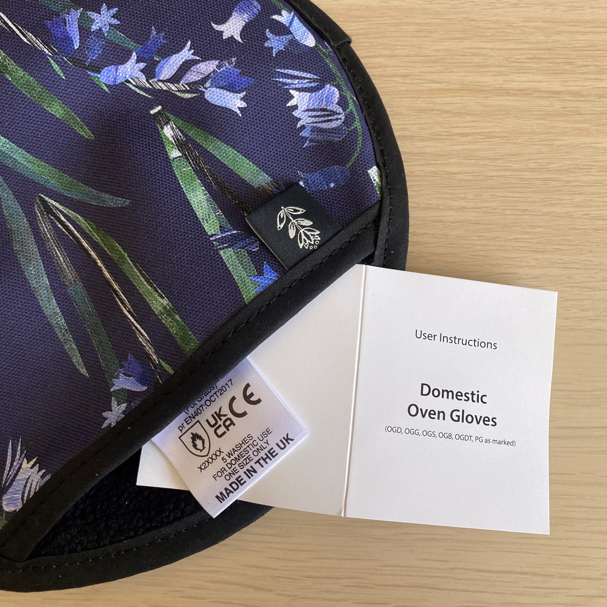 Close up of the Bluebell Single Oven Mitt showing the care label and user instructions.  It has been placed on a wooden table.