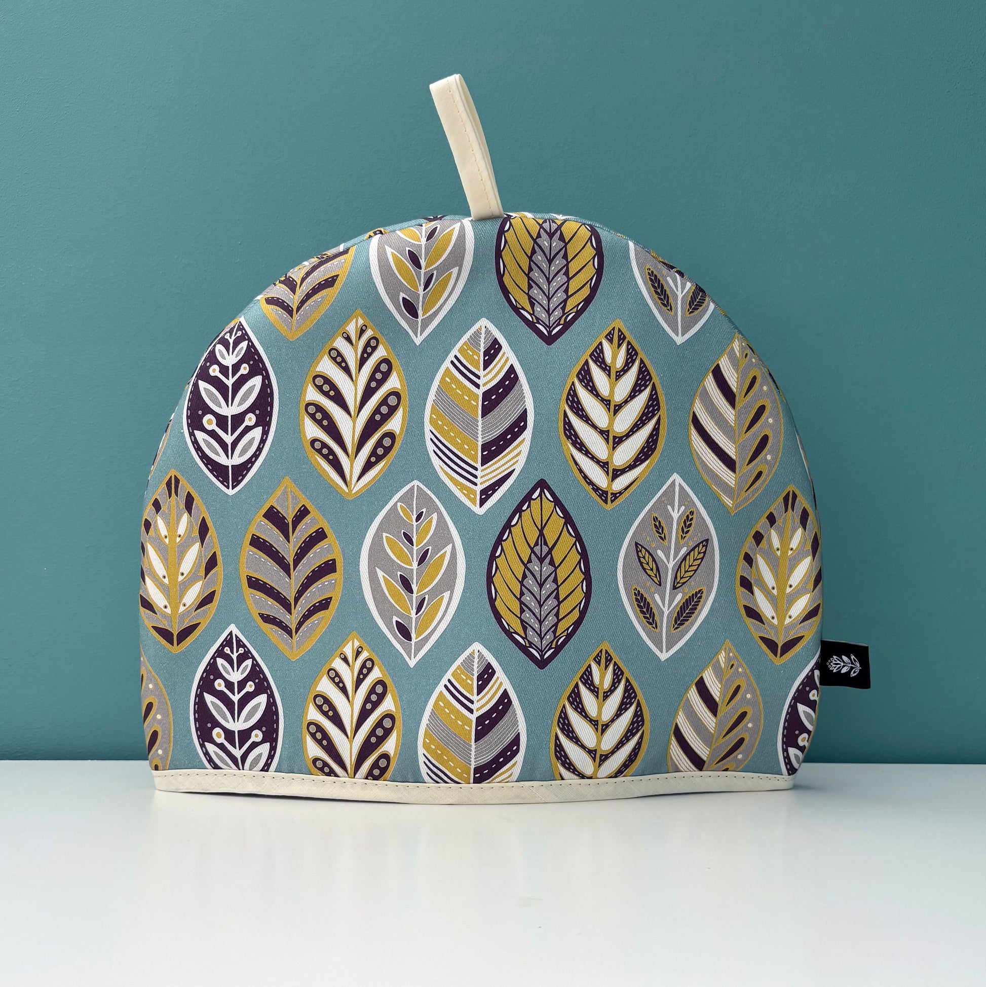 A Blue Leaf Tea Cosy has been placed on a white shelf against a blue green background.