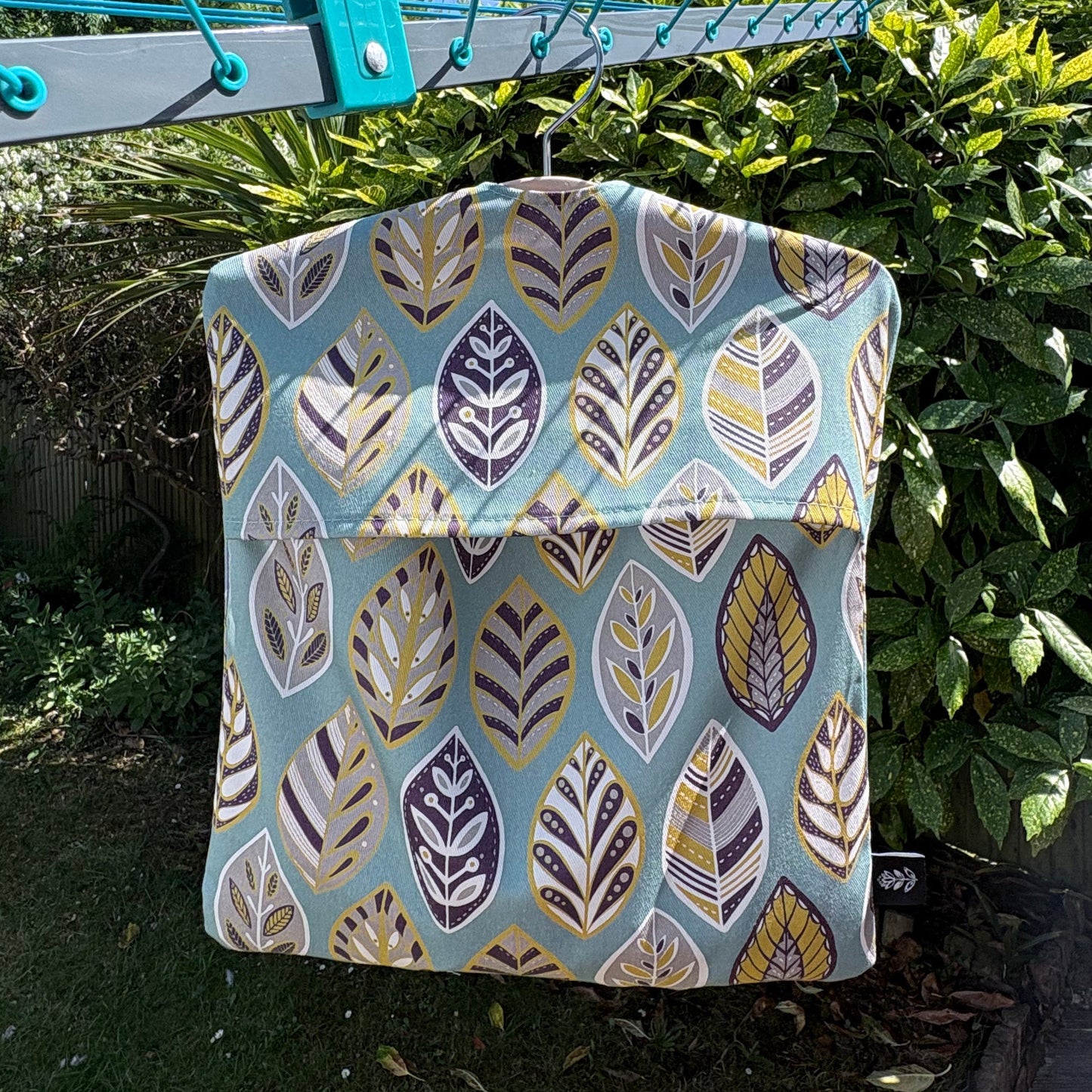A Blue leaf patterned peg bag hanging outdoors on a rotary air drier with greenery.