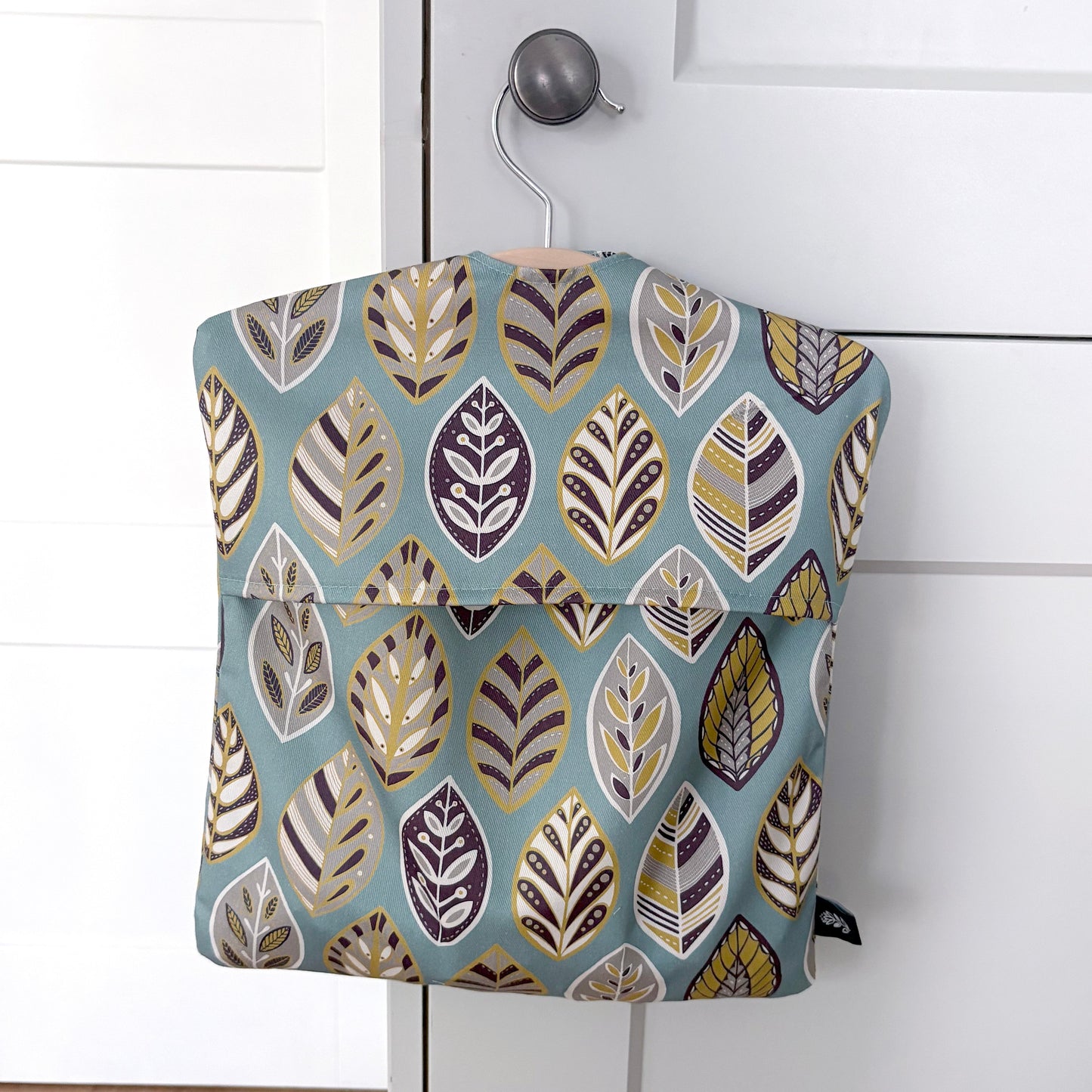 A Blue Leaf peg bag featuring a blue, yellow, grey and off white leaf pattern has been hung on a pale grey kitchen door knob.