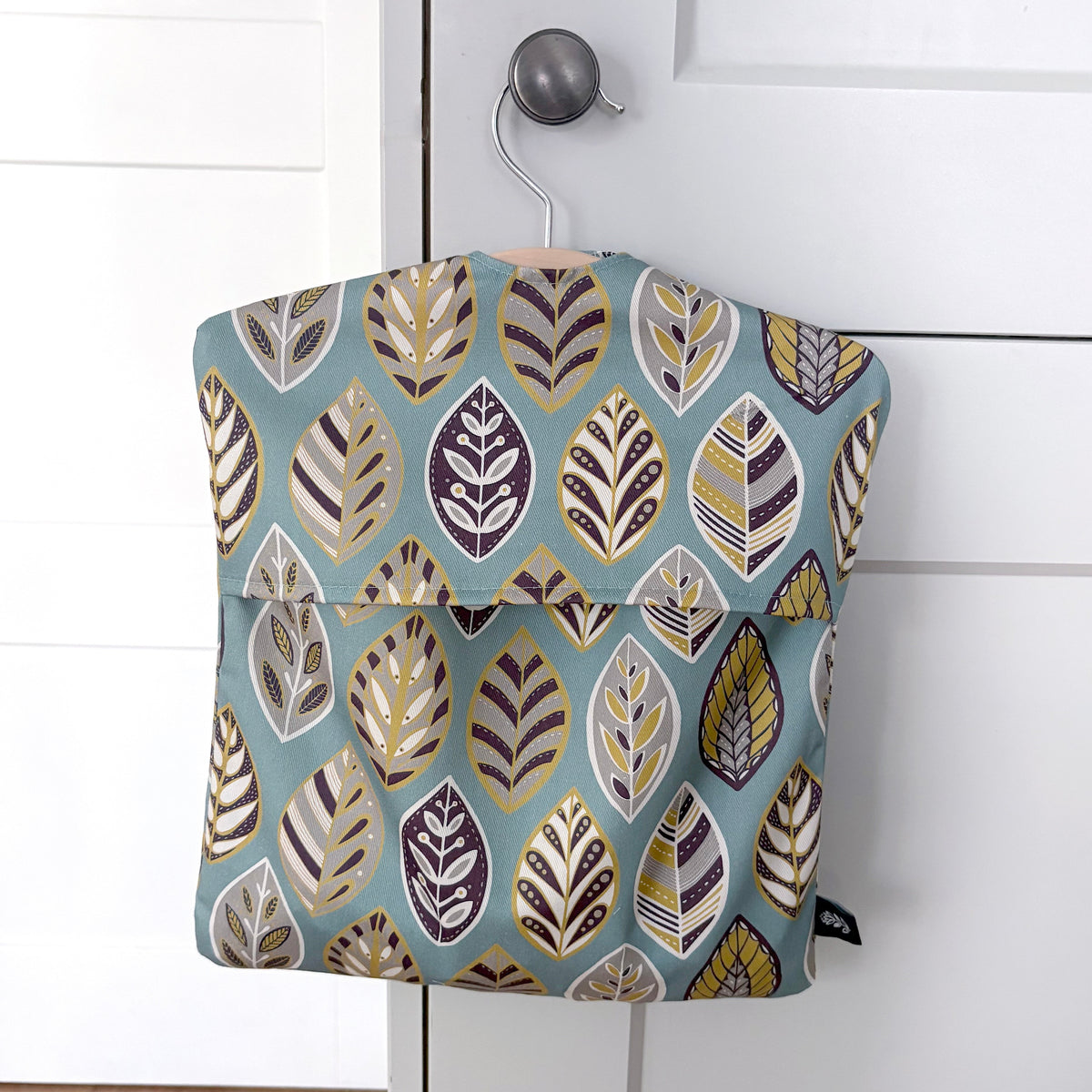 A Blue Leaf peg bag featuring a blue, yellow, grey and off white leaf pattern has been hung on a pale grey kitchen door knob.