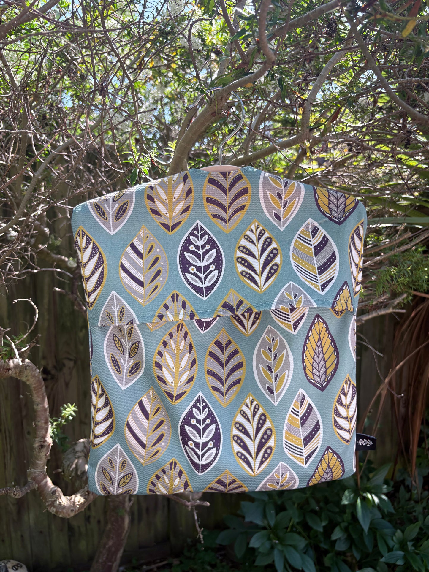 A Blue Leaf patterned peg bag hanging outdoors on a wooden fence with greenery.