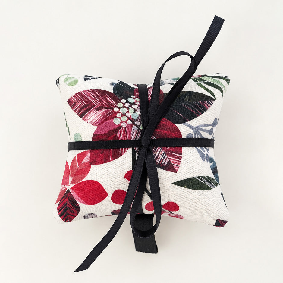 A pack of three square lavender bags on a winter floral pattern on a white base with a white background and a red, green, and black floral pattern, tied with a black ribbon bow in the centre, on a plain off-white background.