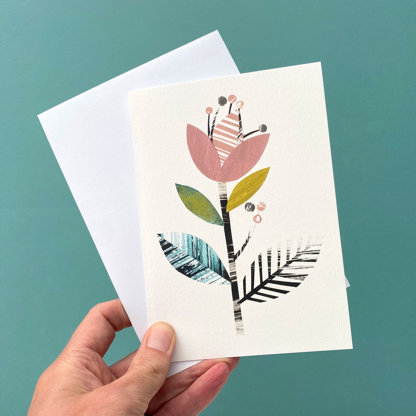 A Card featuring a stylised floral motif in Dusky Pink, green, yellow and black on a white ground, is being held with a white envelope against a blue green background.