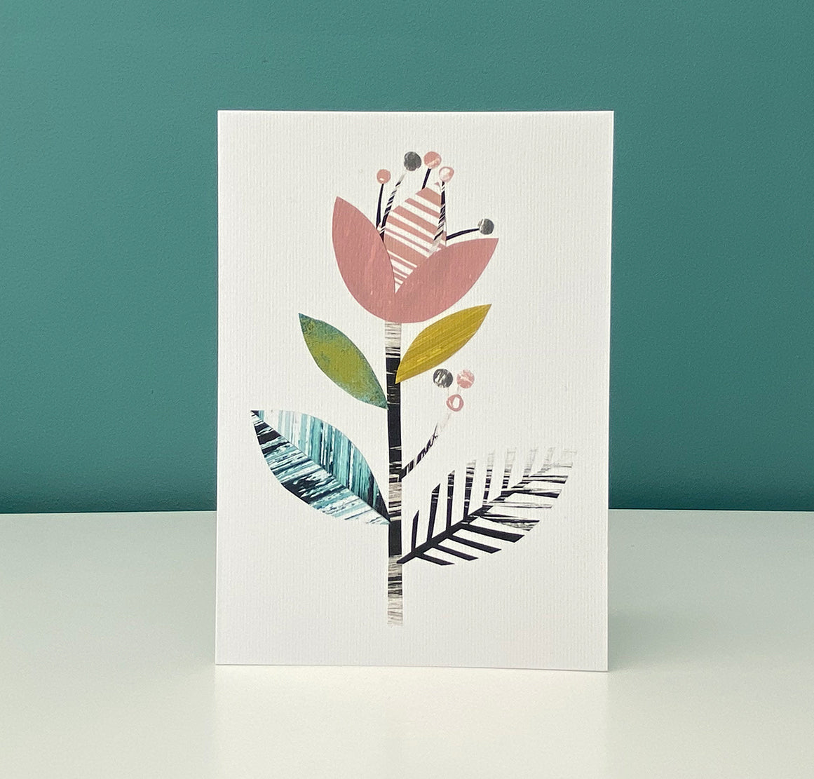 A Card featuring a stylised floral motif in Dusky Pink, green, yellow and black on a white ground has been stood on a white shelf with a blue green background.