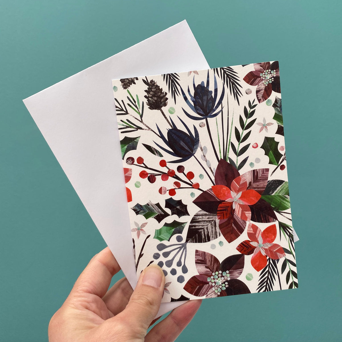 A Winter Floral Christmas card is being held with a white envelope against a blue green wall.