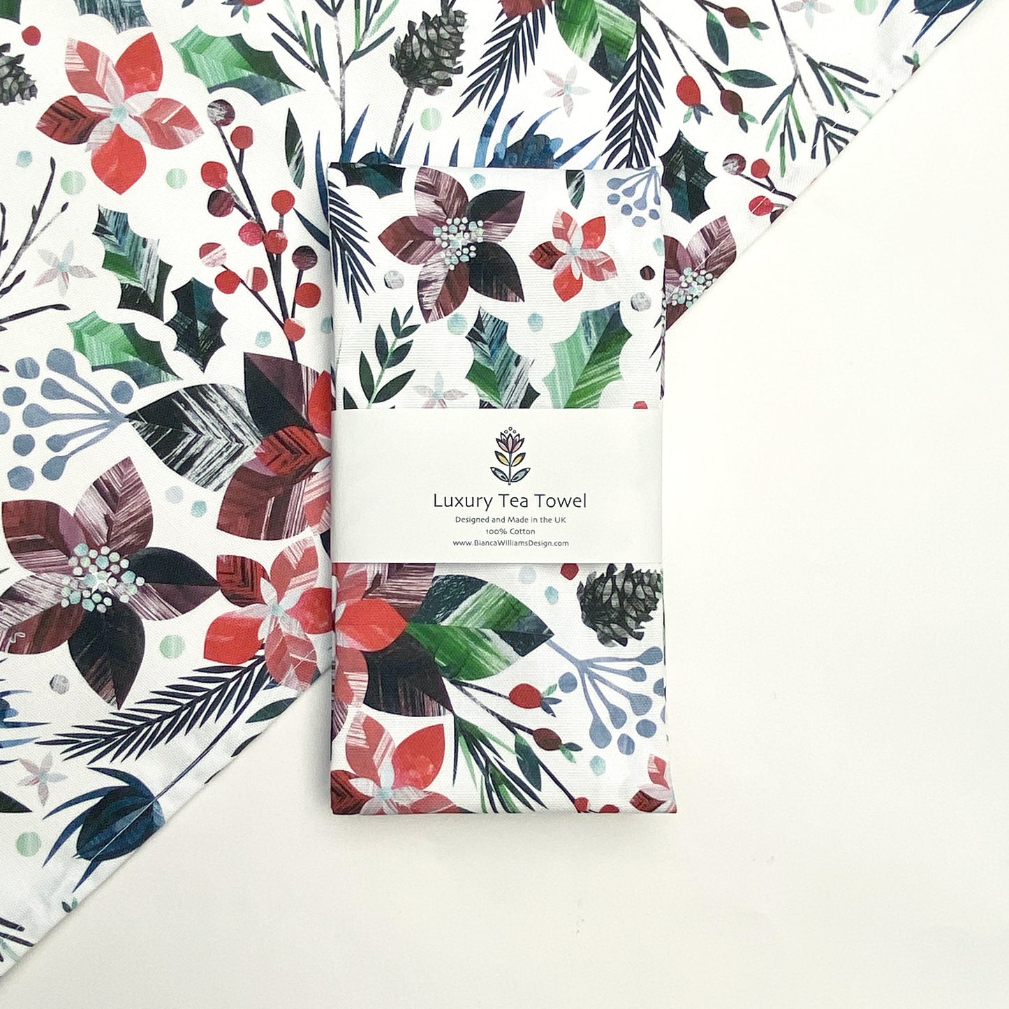 A Winter Floral Tea Towel has been packaged in a branded belly band and has been placed on a white background which also has an opened out winter floral tea towel also placed at an angle behind it.