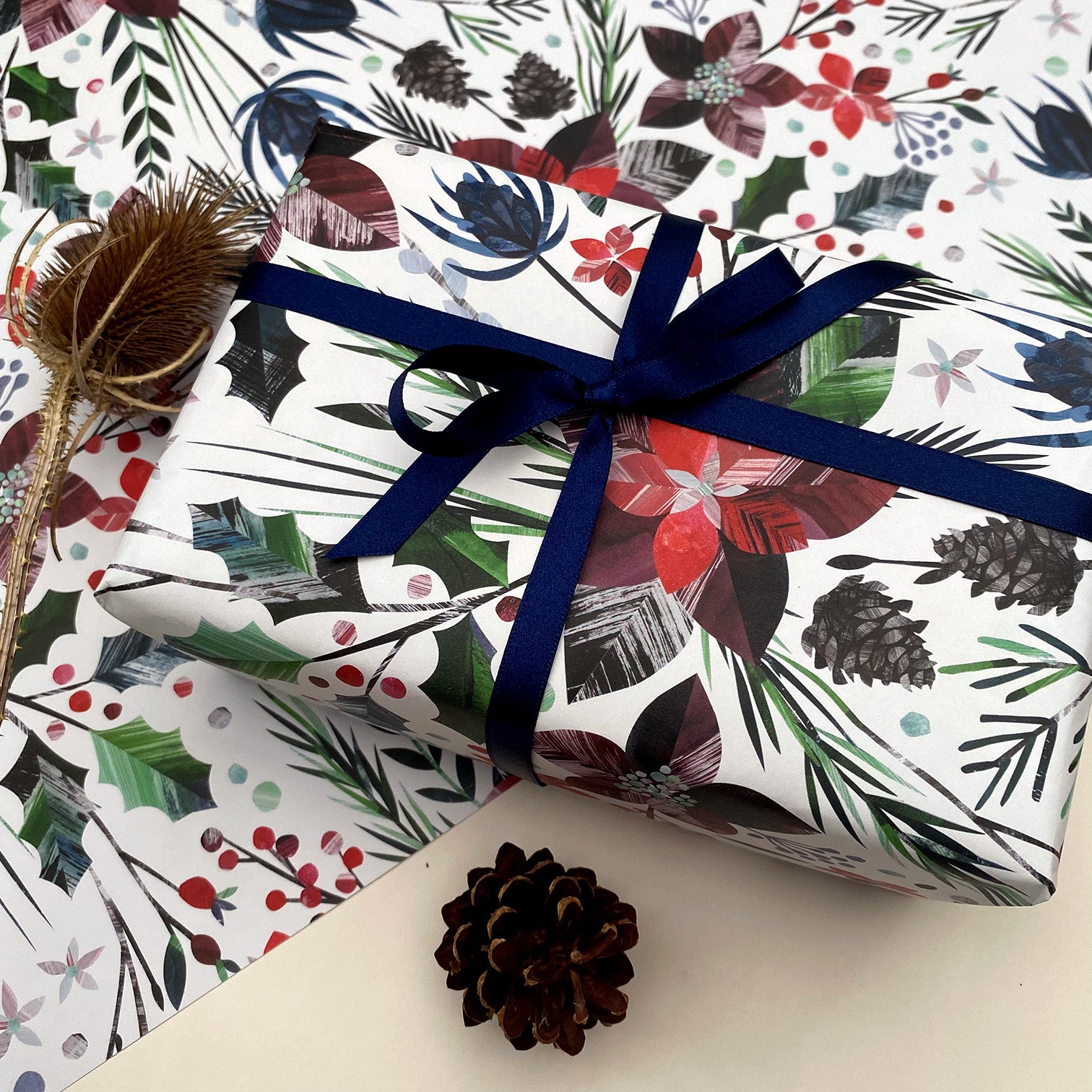 A present has been wrapped in the winter Floral Christmas wrapping paper and has been tied with dark blue satin ribbon in a bow.The present has been placed on a sheet of the winter floral gift wrap.  A thistle and pinecone can also be seen.