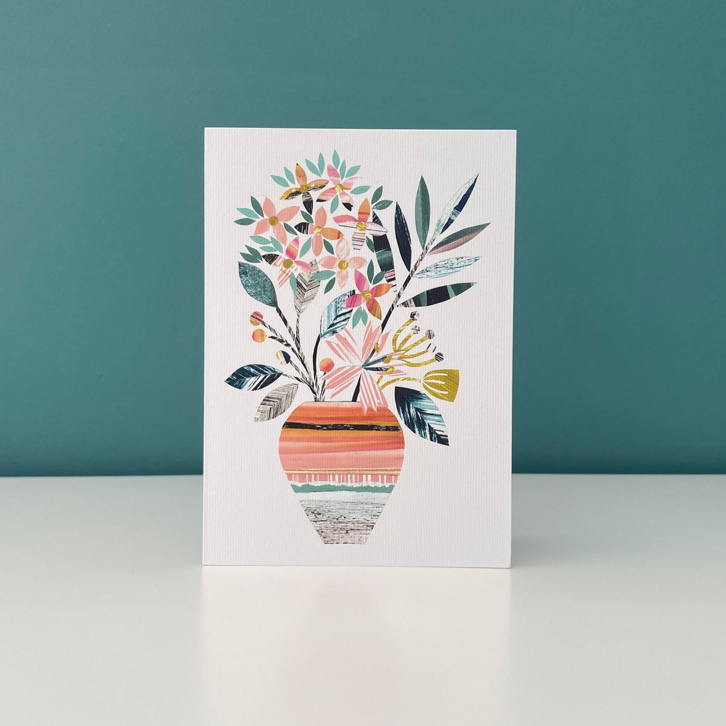 A white greeting card with a print of a colourful textured vase of flowers in green, orange, yellow and pinks has been stood up on a white shelf and has a blue green wall behind it.