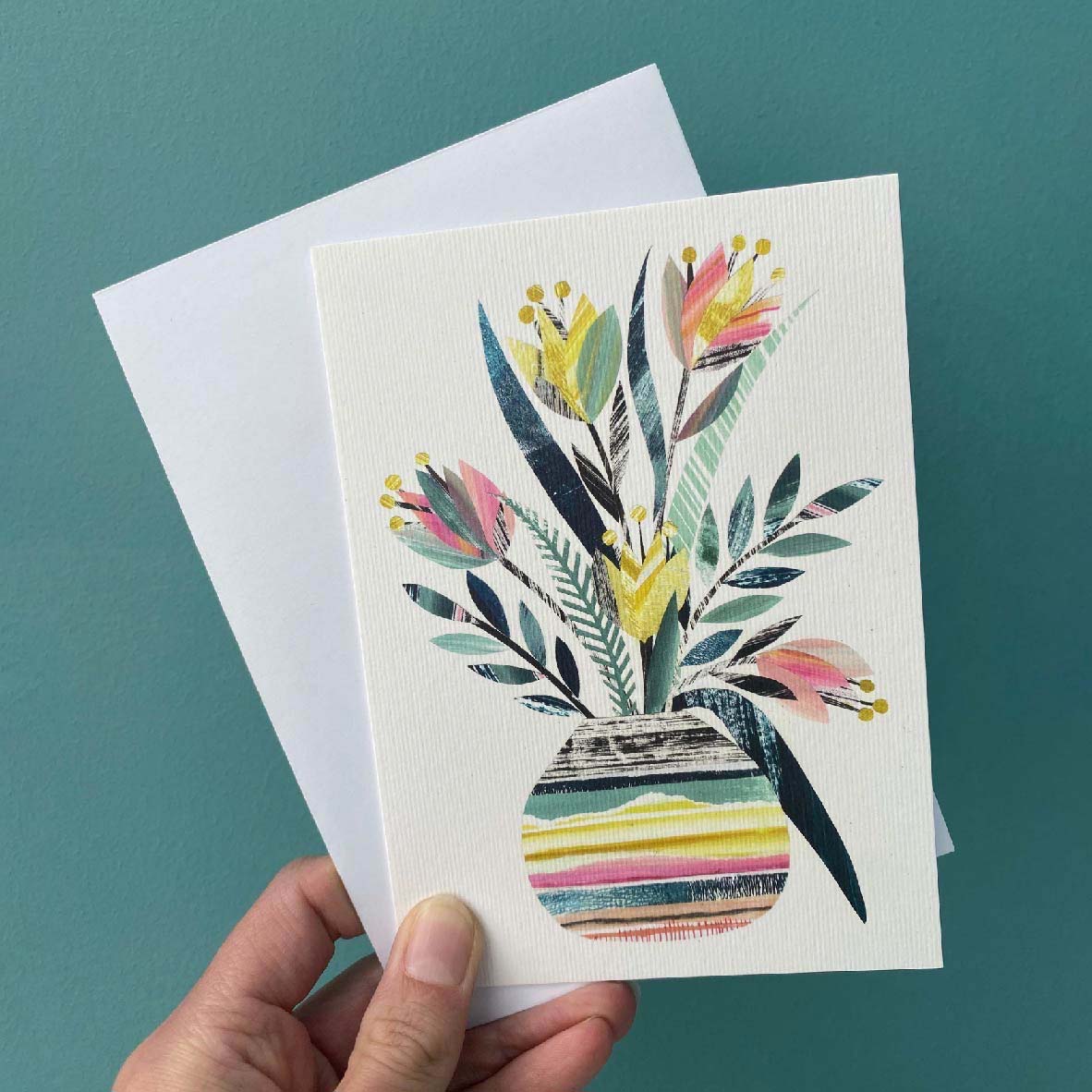 A greeting card featuring a colourful vase of stylised Tulips in yellows, pinks and green on a white card, is being held with a white envelope against a blue green background.