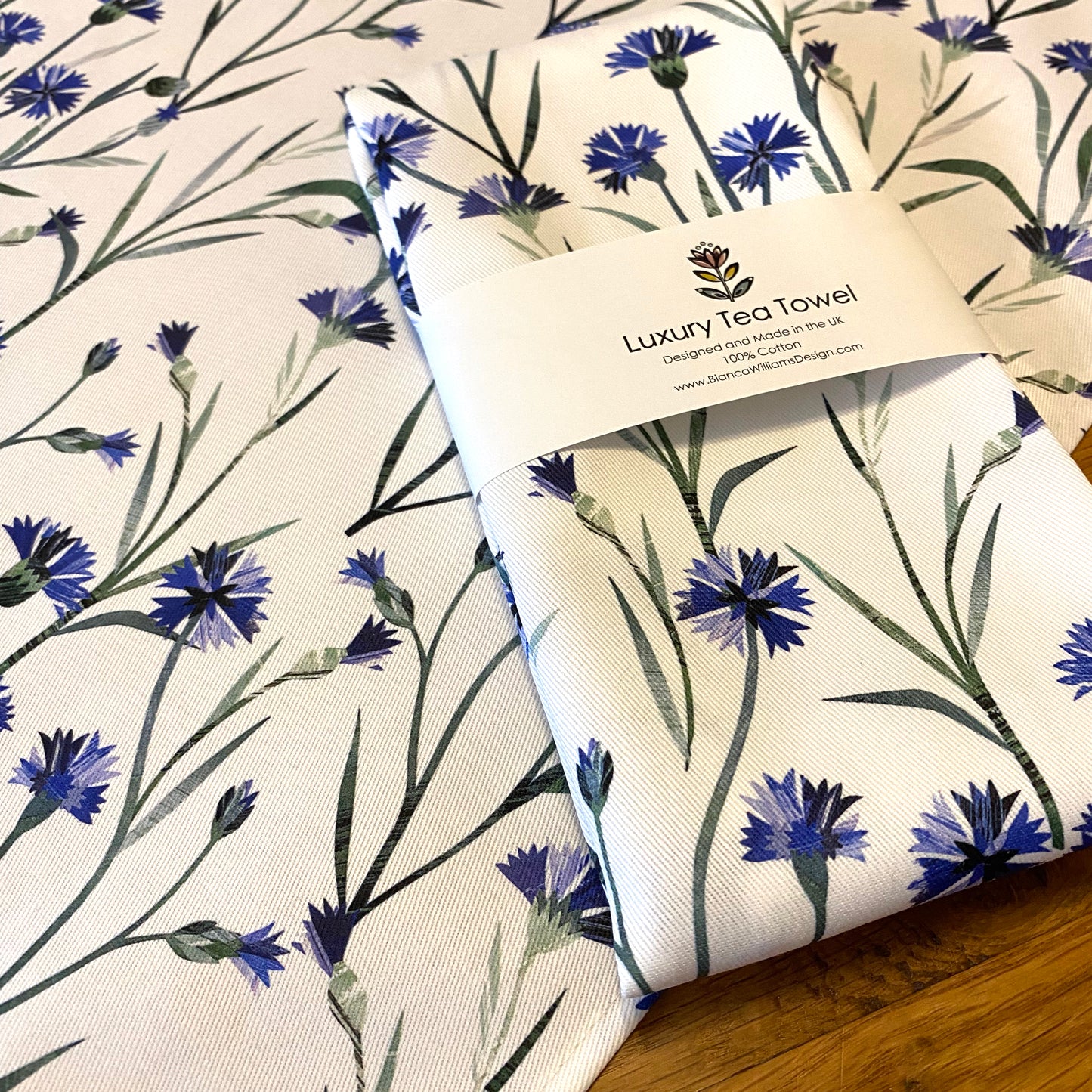 A packaged cornflower tea towel has been placed on a wooden table with an opened out Cornflower Tea Towel.