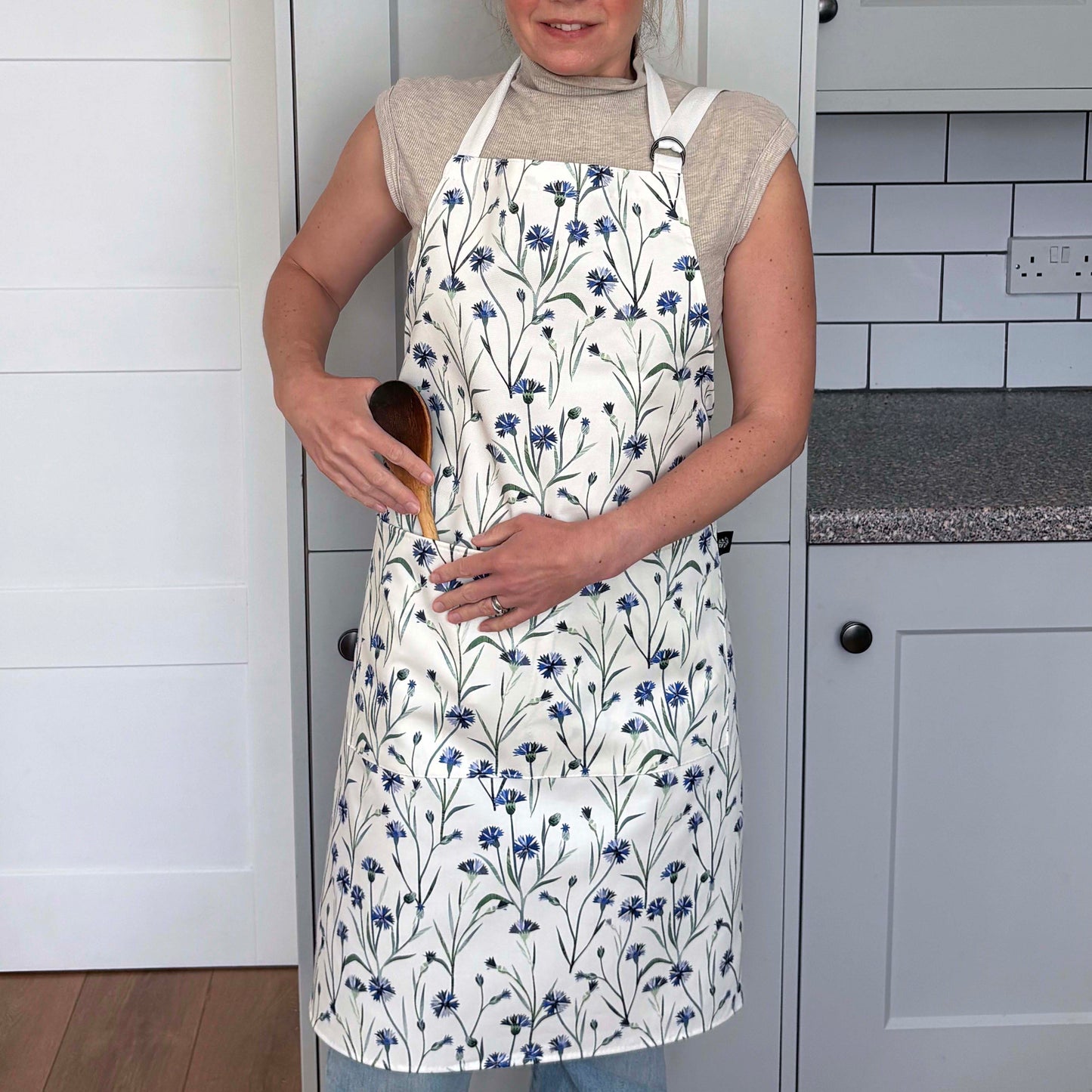 A lady is wearing the Cornflower apron with two pockets on the front and an adjustable neck strap in a grey kitchen.  She has tied the apron around her waist.