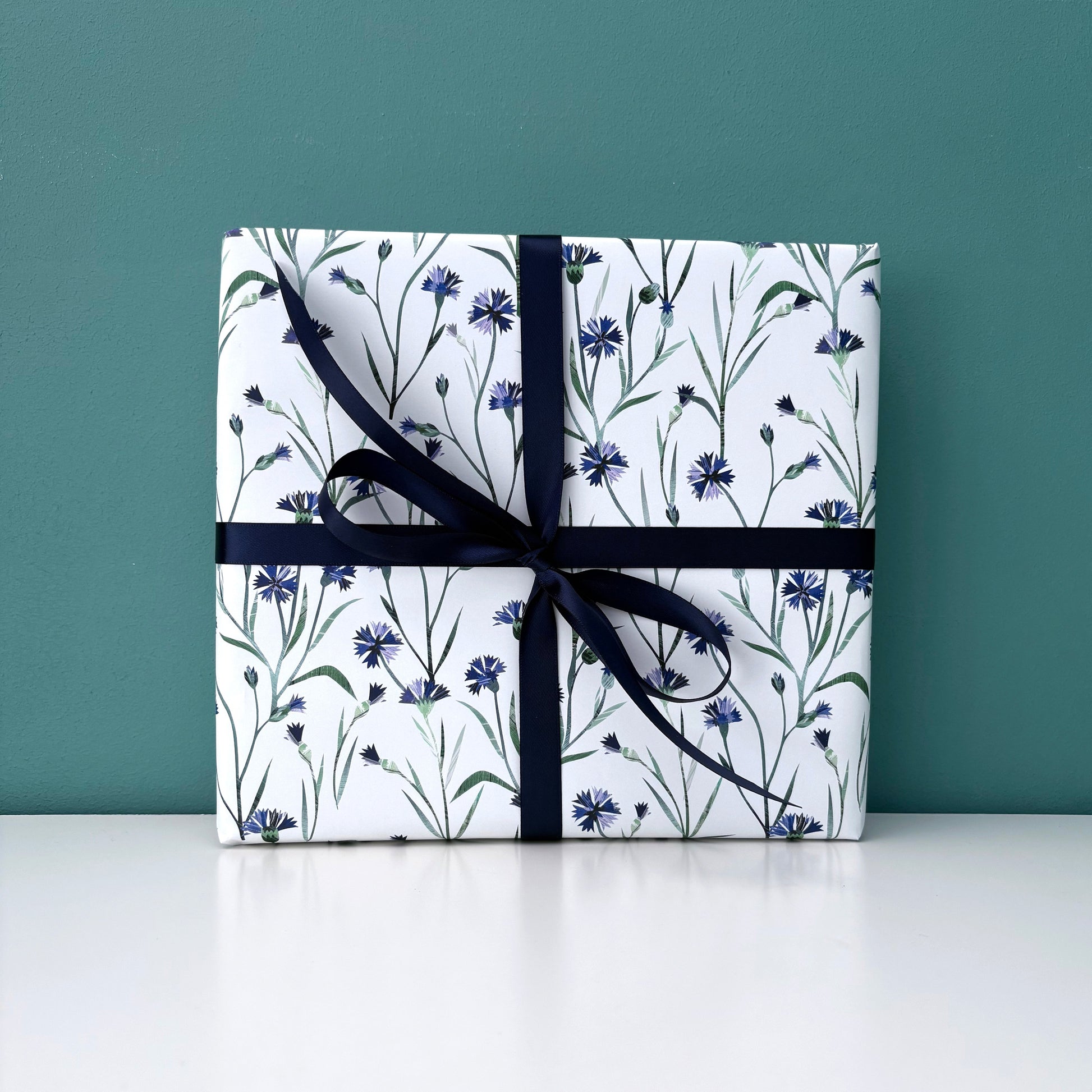 A present has been wrapped in Cornflower Gift Wrap and has been tied with a coordinating Navy satin ribbon and placed on a white shelf against a blue/green backdrop.