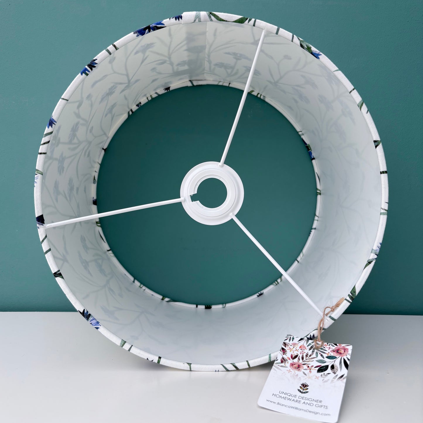 Inside of a medium Cornflower lampshade, showing the white inside.  It has been placed on a white shelf against a blue green wall.