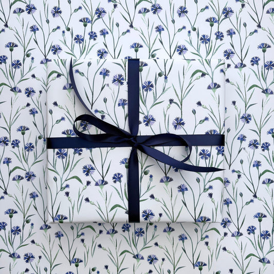 A present has been wrapped in the Cornflower Gift Wrap and has been tied with a coordinating Navy satin ribbon and placed on a sheet of laid out Cornflower gift wrap. The gift wrap features pretty Blue Cornflowers with green leaves on a off white background.