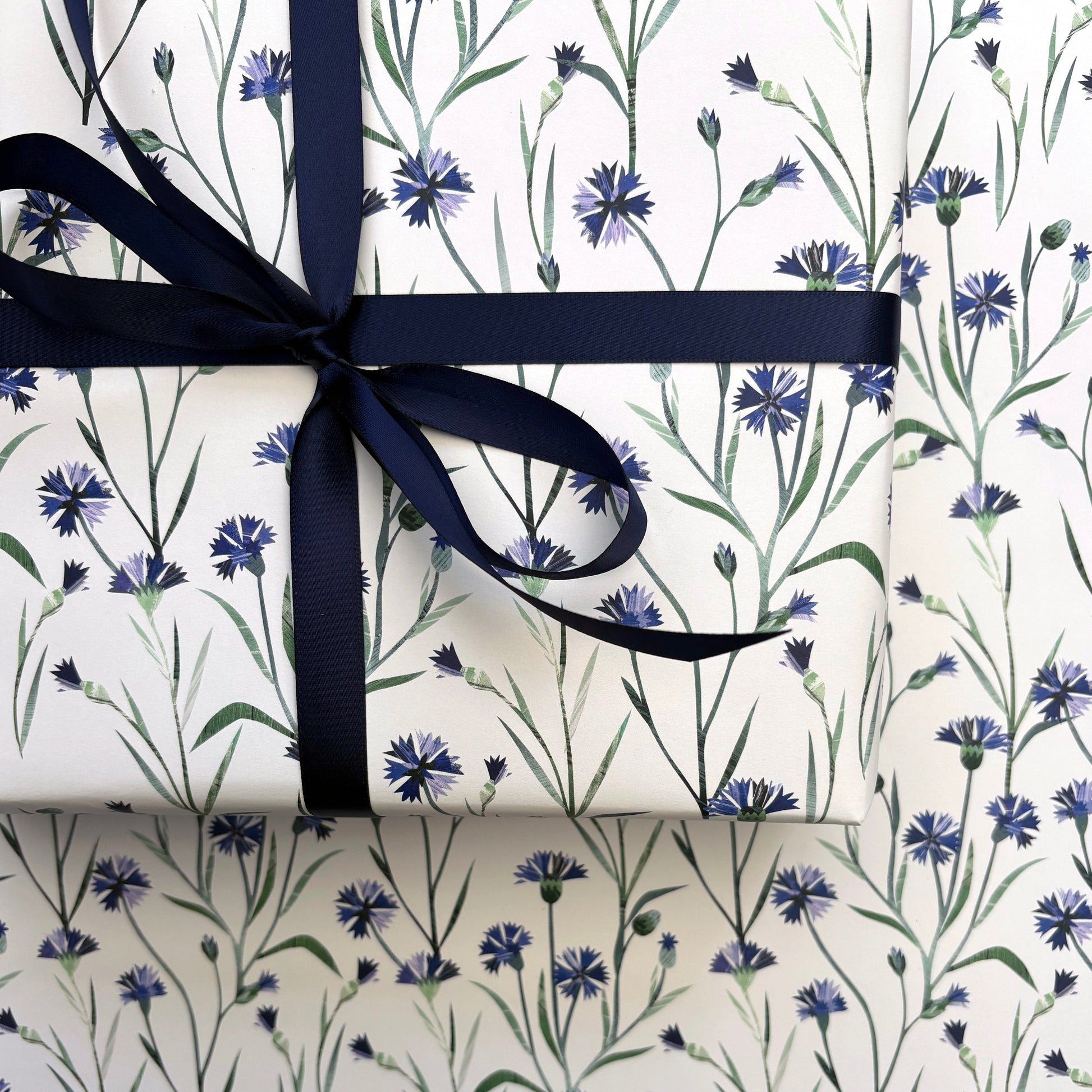 A close up of a present that has been wrapped in Cornflower Gift Wrap and tied with a coordinating Navy satin ribbon and placed on a sheet of laid out Cornflower gift wrap.