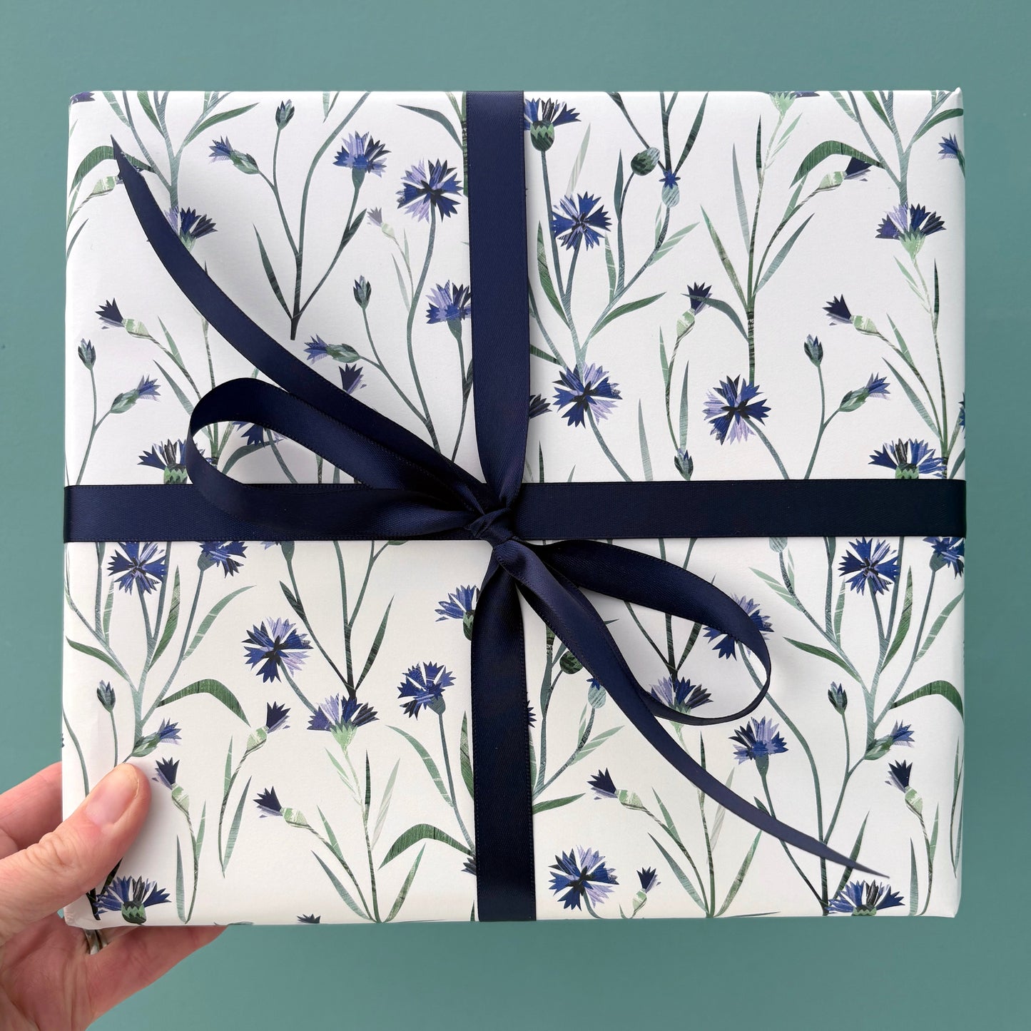 A present has been wrapped in Cornflower Gift Wrap and has been tied with a coordinating Navy satin ribbon and is being held against a blue/green backdrop.