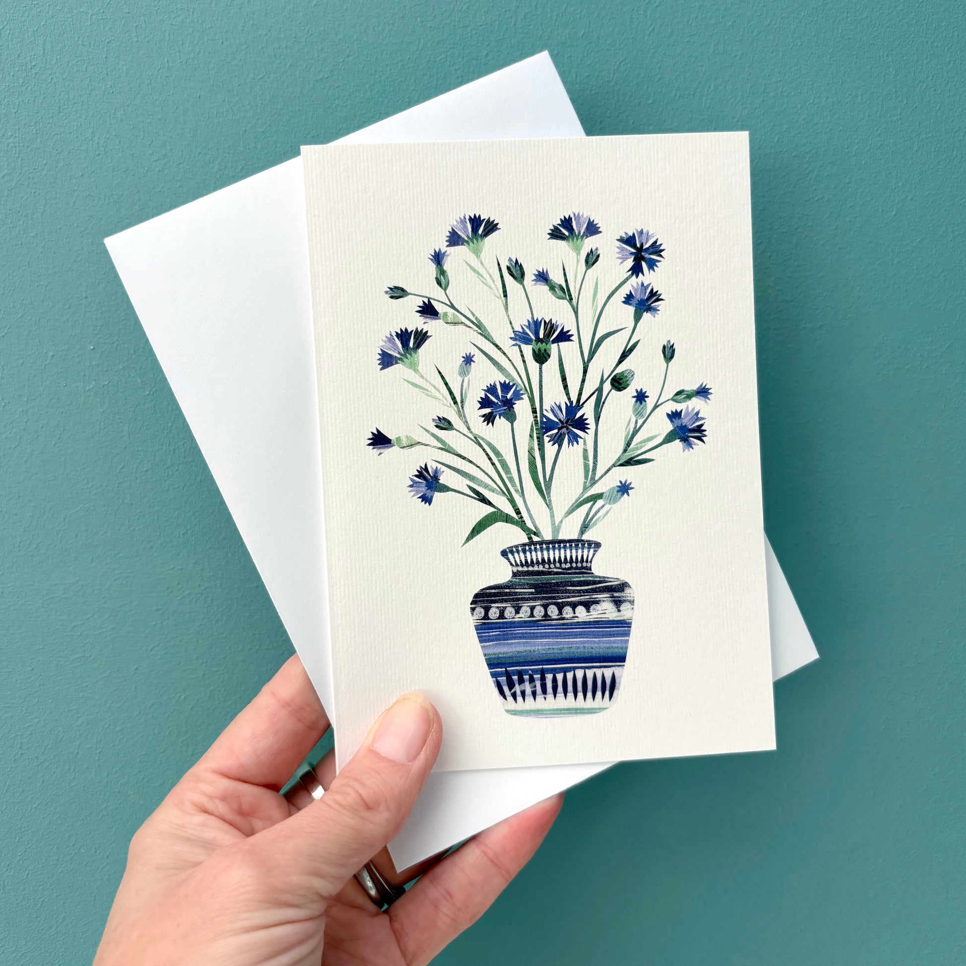 Greeting card with Blue Cornflowers in a Vase on a white card held with a white envelope against a teal background.