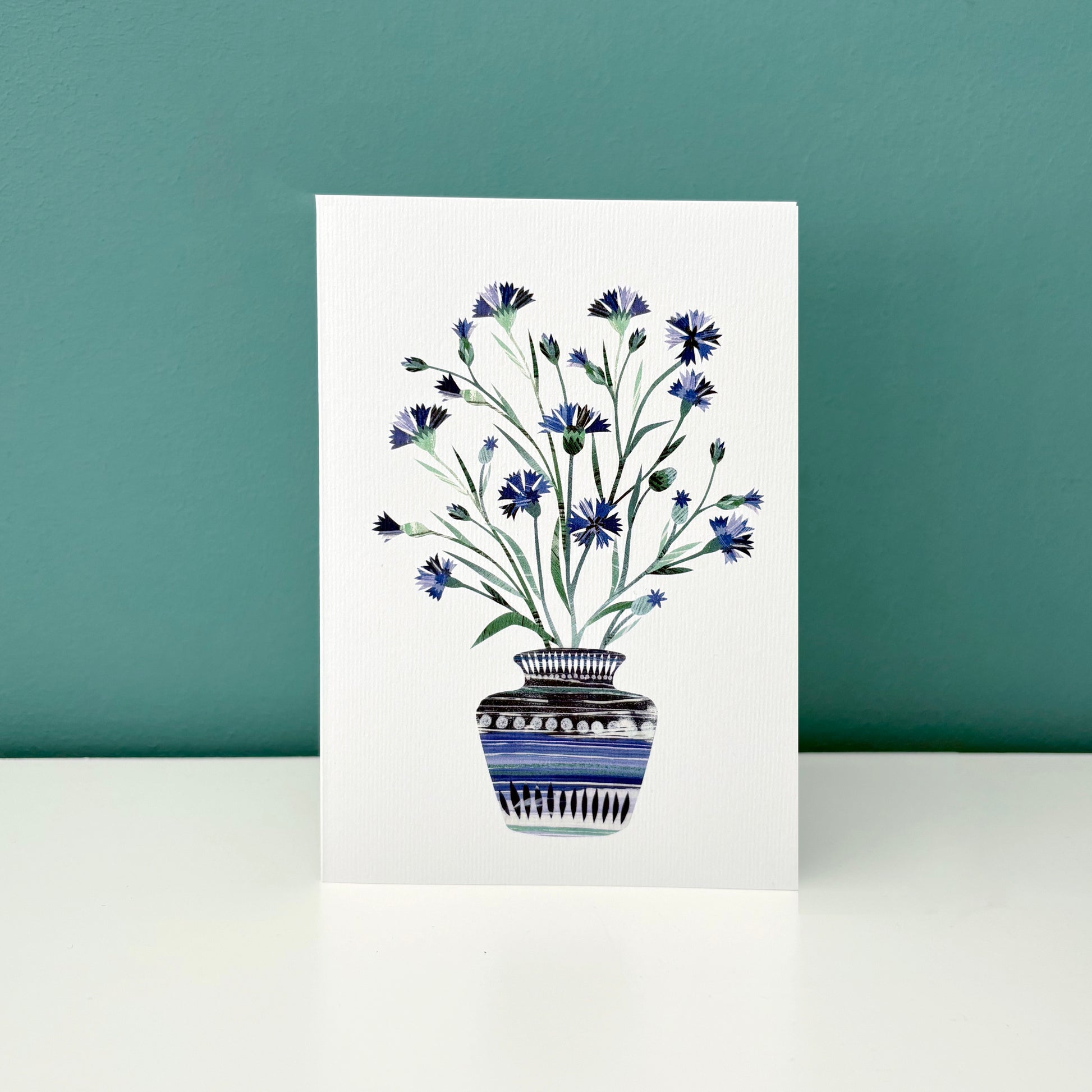 A white card with a Vase of Blue Cornflowers on it.  The Card has been stood on a white shelf with a blue green wall behind it.