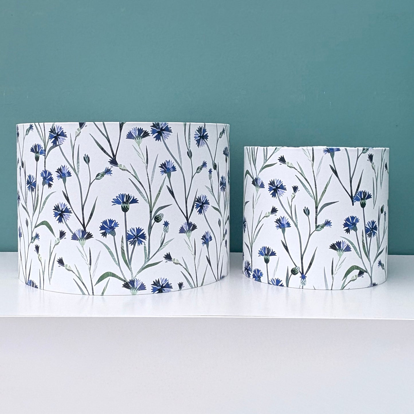A medium and a small floral lampshade have been placed side by side on a white shelf against  a blue green wall.  The lampshades have a blue cornflower pattern with green leaves on a white background.