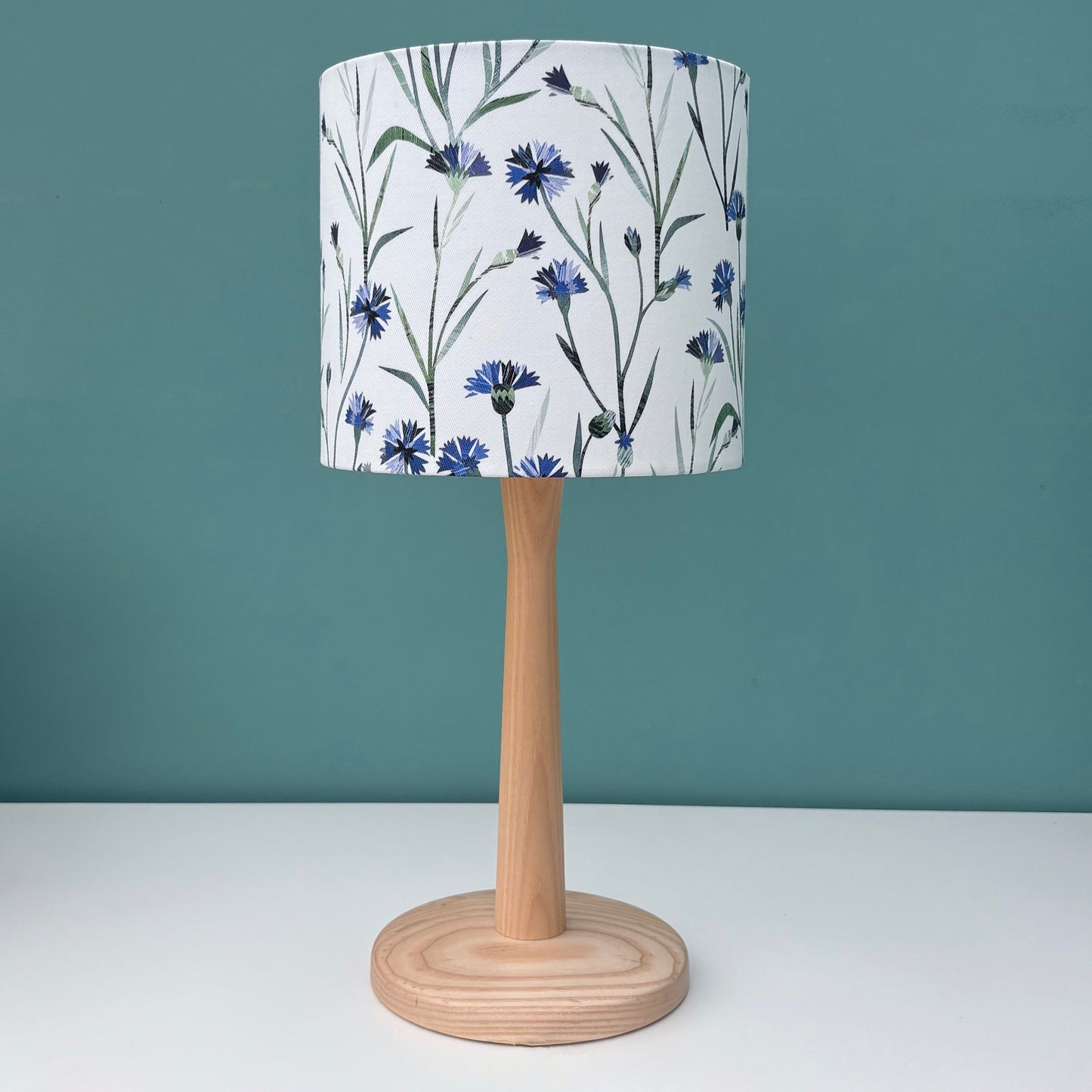 A small Floral lampshade featuring blue Cornflowers with green leaves on a white background has been placed on a wooden table lamp.  The table lamp has been put on a white shelf against a blue green wall.