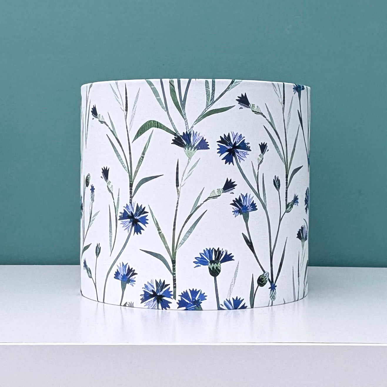 A small floral lampshade featuring blue cornflowers with green leaves on a white background has been placed on a white shelf with a blue green background.