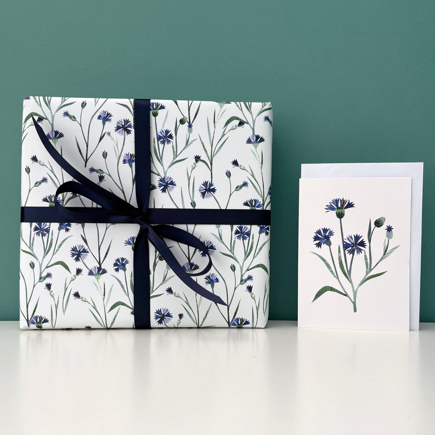 A present wrapped in Cornflower wrapping paper and tied with coordinating Dark blue satin ribbon has been placed next to a matching white card featuring a matching Cornflower on it.  Both the present and the card have been placed on a white shelf with a blue green background.