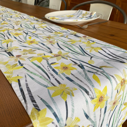 A close up of a daffodil Table runner featuring all the colours, textures and tones of these bright yellow and orange flowers with green leaves, printed onto a white cotton background.