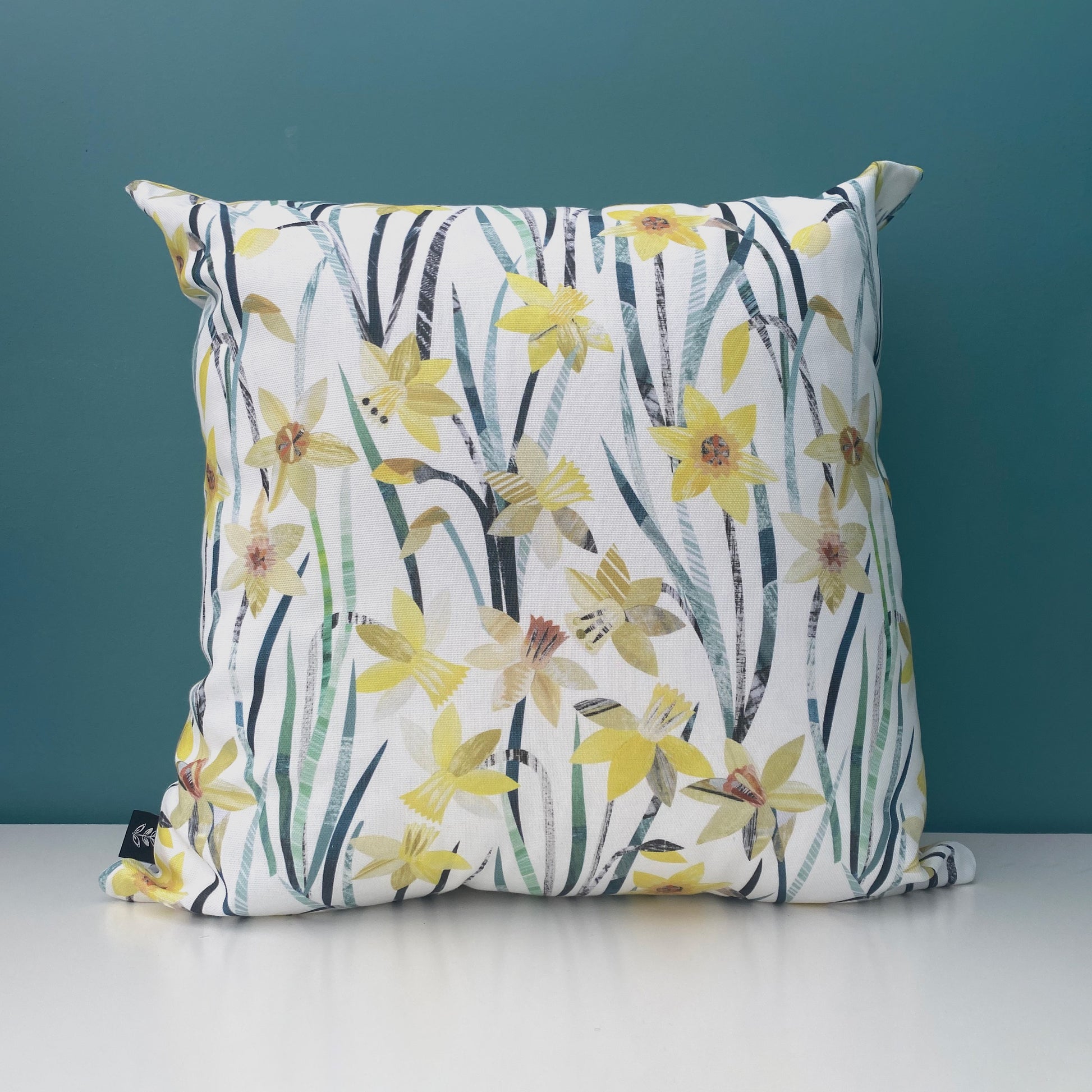 A Daffodil Cushion featuring pretty textured Yellow Daffodil Flowers with green leaves has been printed onto a white background.  The cushion has been placed onto a white shelf with a blue/green wall behind.