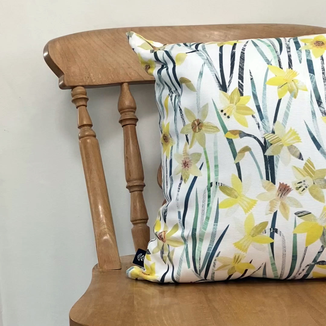 A Daffodil Cushion has been placed onto a wooden Dining chair against a pale grey wall.