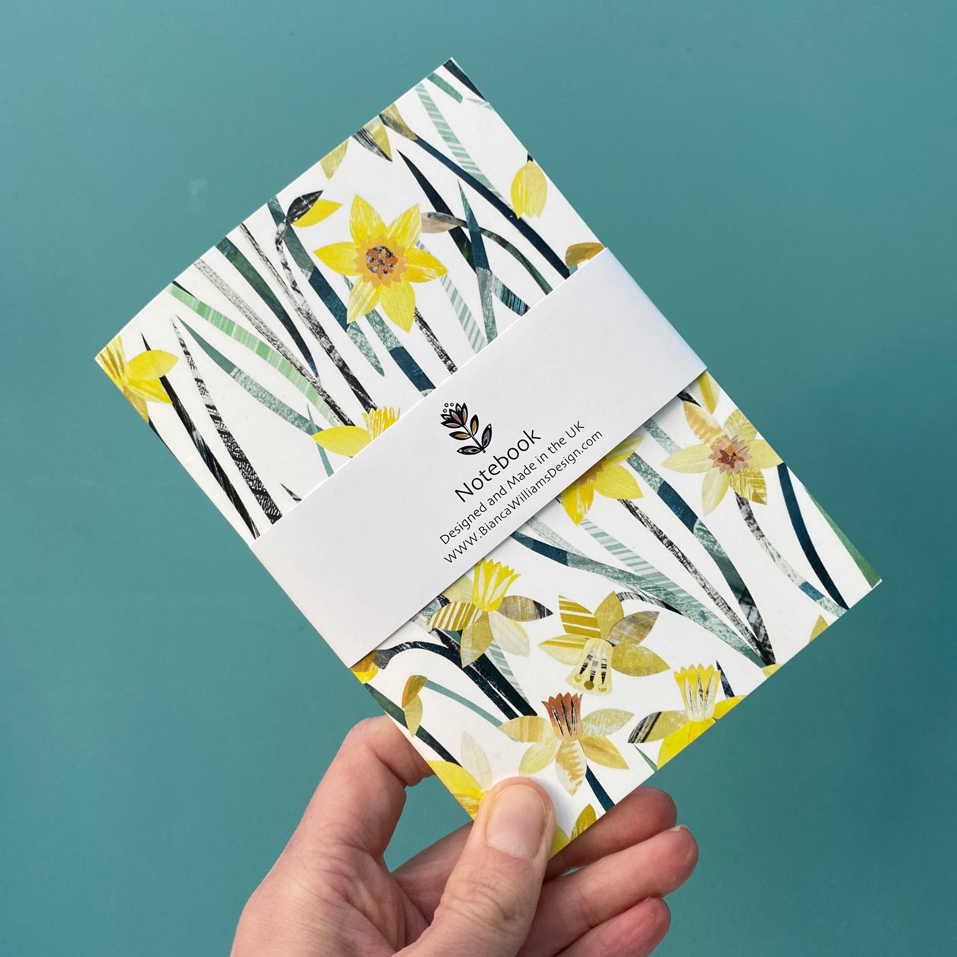 Small white notebook with all over Daffodil pattern packaged in a white branded belly band and held in front of a blue green back ground.