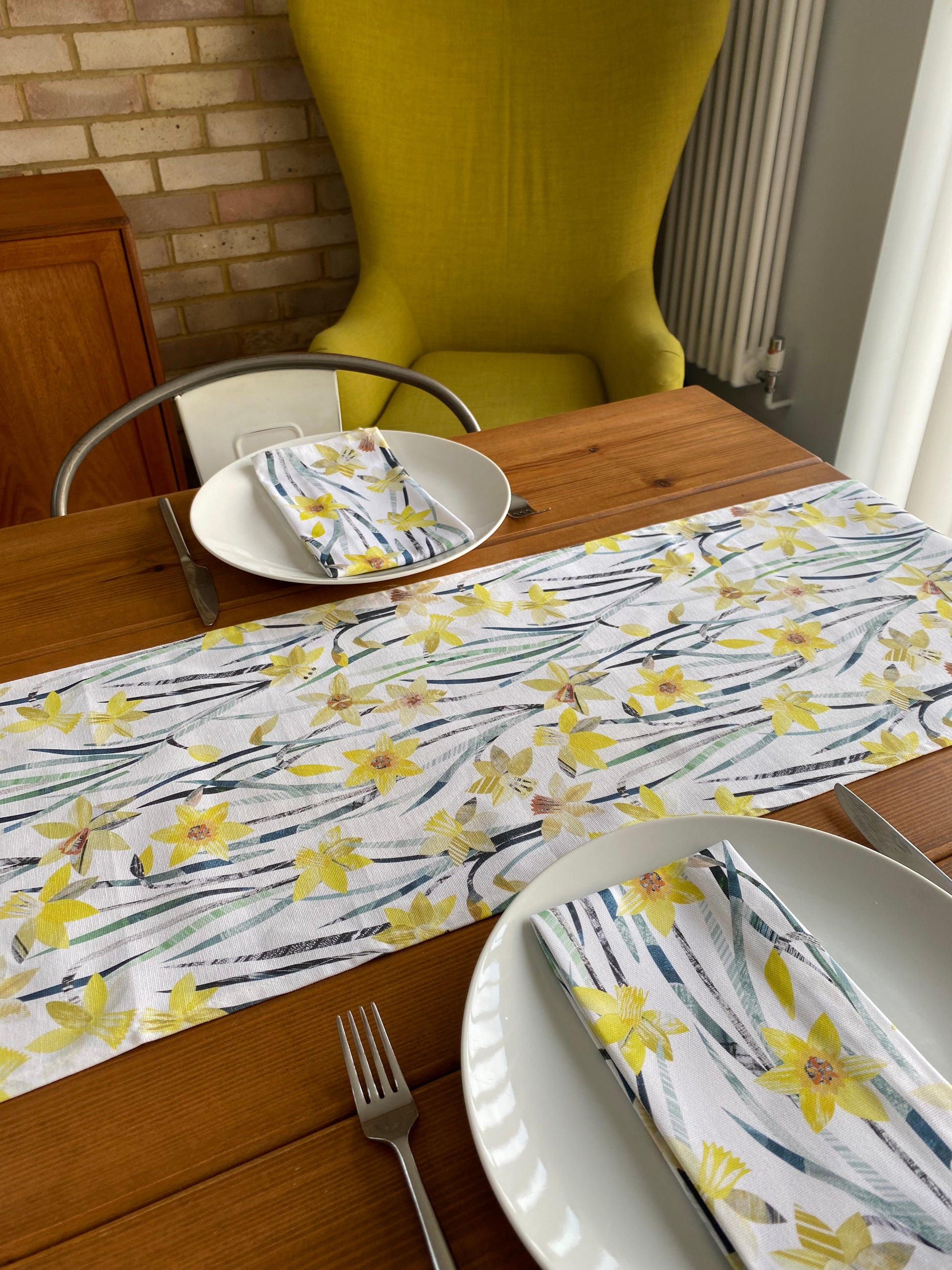 A Daffodil Table runner has been laid down the centre of a wooden table.  The table has been set for two, with two white plates and matching daffodil Napkins.