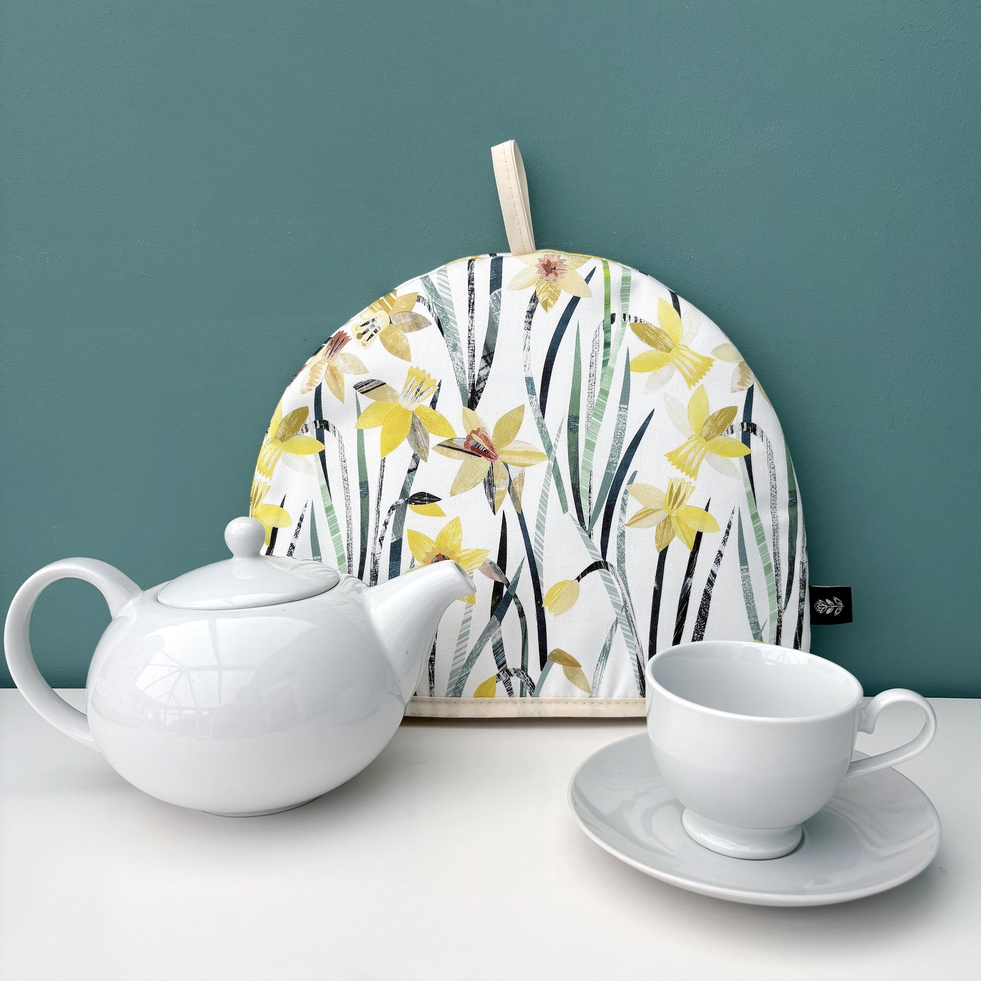 A Daffodil Tea Cosy and a white Teapot, cup, and saucer on a white surface against a teal wall.
