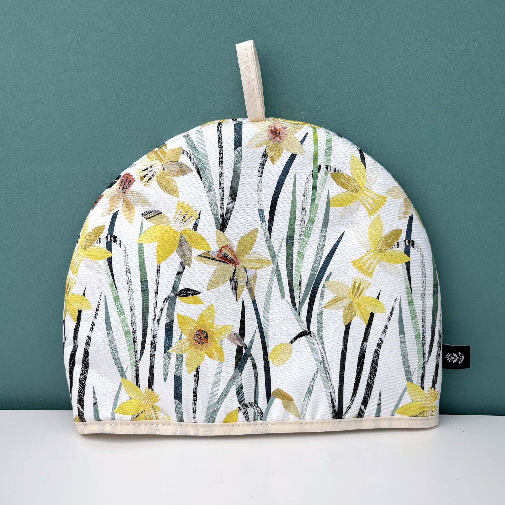 Daffodil Tea cosy with a yellow spring floral design on a plain background, has been placed on a white shelf with a teal background.