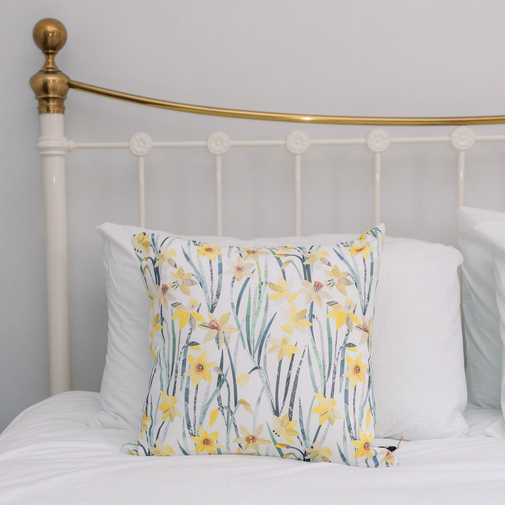 A decorative square throw pillow with a yellow daffodil floral print, styled on a white bed with a white metal headboard featuring a gold accent.