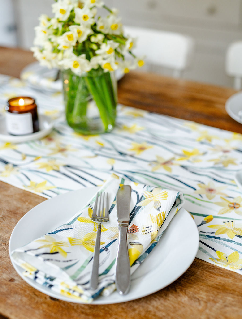 Dinner setting with Daffodil floral table runner and matching napkins, plates, cutlery, and a vase of flowers.